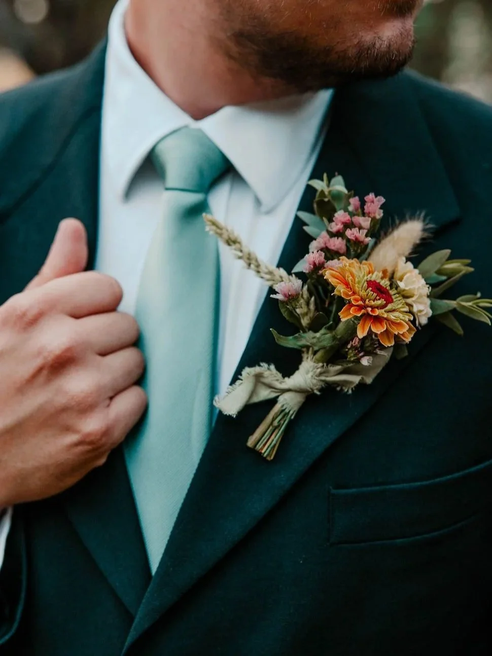 Close-up of a man dressed in a dark suit, white shirt, and green tie, with a floral boutonniere pinned to his lapel.