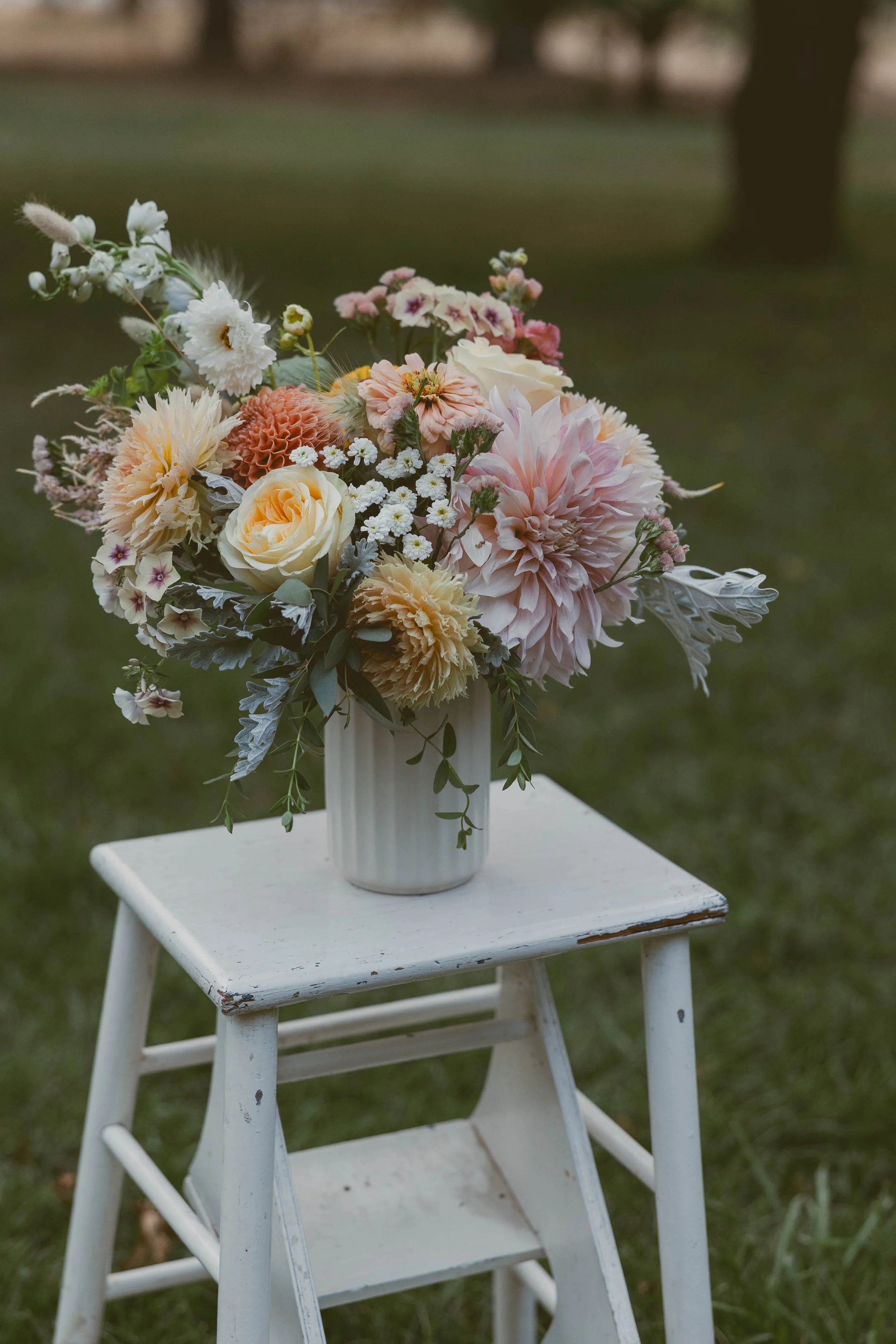 A bouquet of pastel flowers in a white vase placed on a small, rustic white wooden stool outdoors.