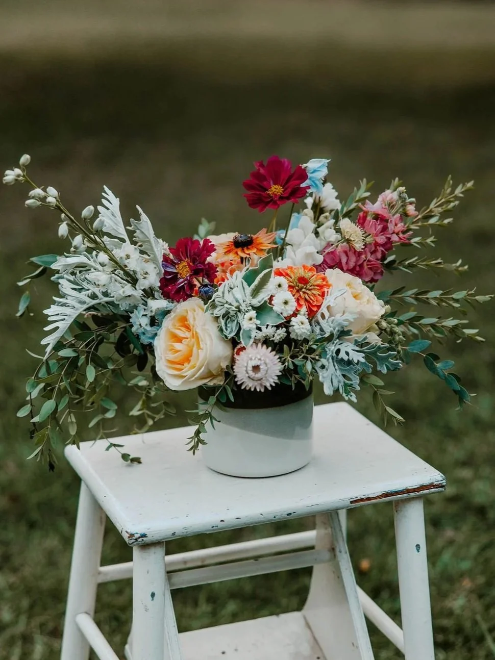 A colorful bouquet of flowers in a white and green vase placed on a small white wooden stool outdoors.