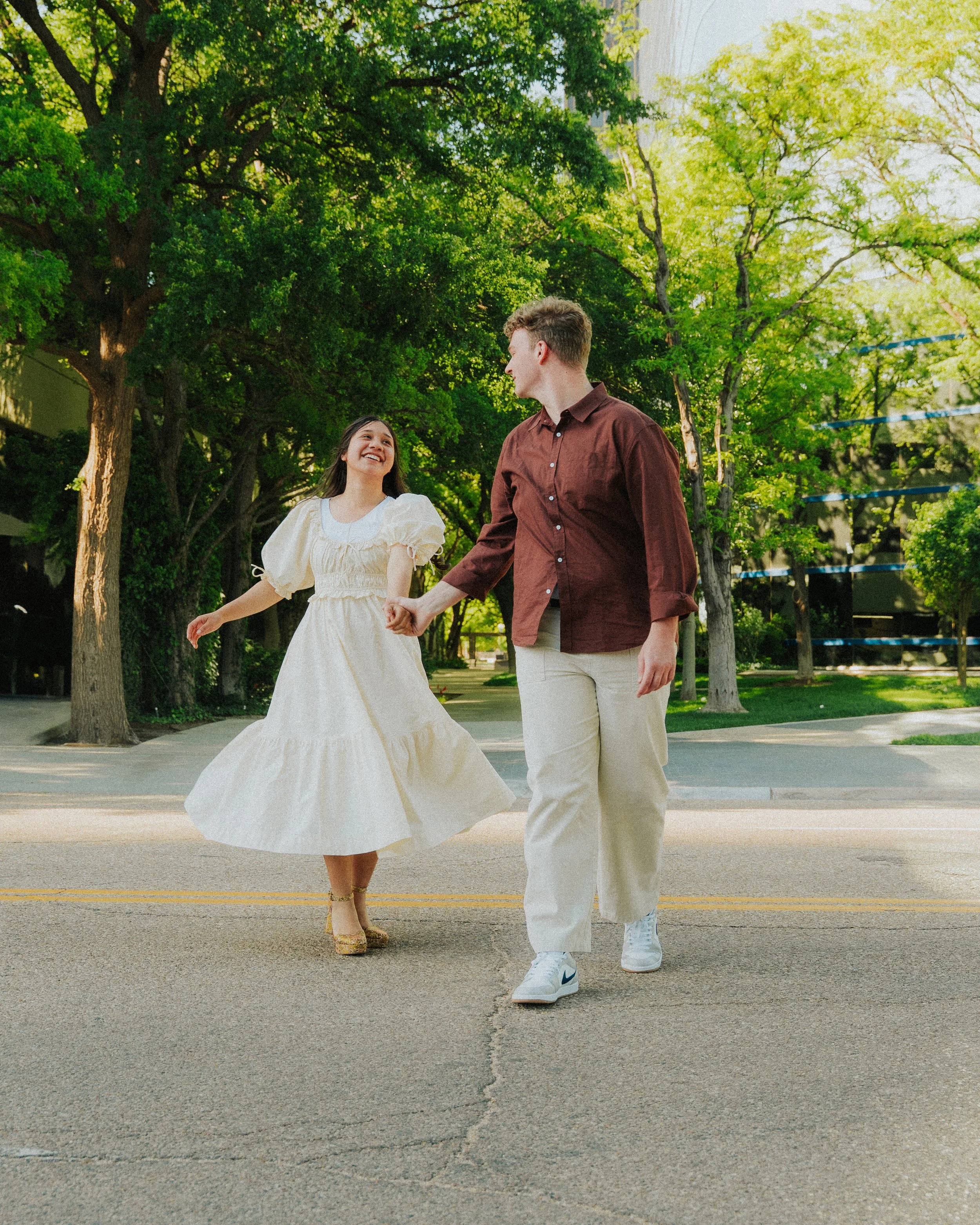A young woman in a white dress and gold heels and a young man in a maroon shirt and beige pants holding hands and dancing on a street with green trees in the background.