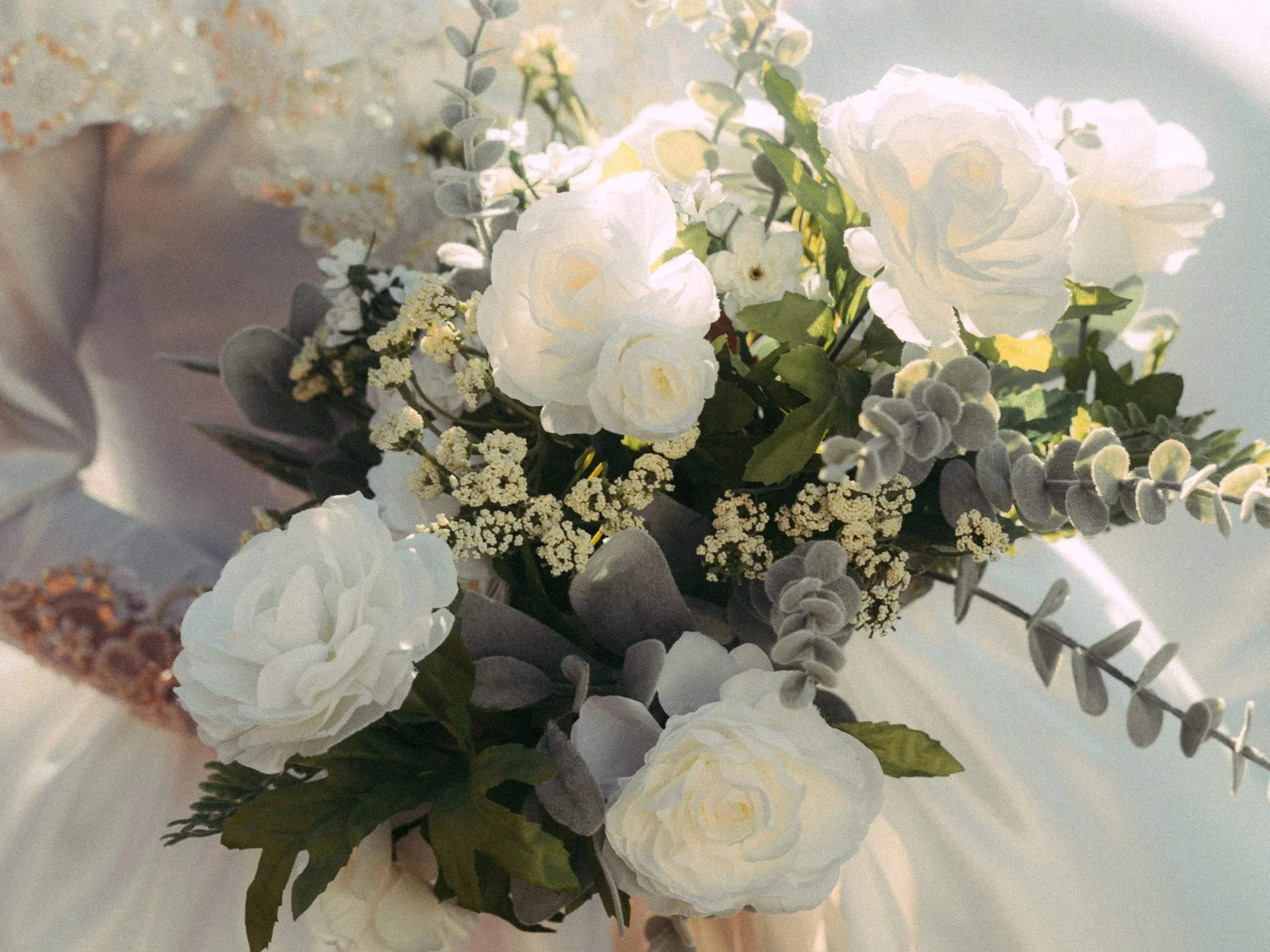 A bouquet of white roses, small white flowers, and greenery on a soft fabric background.