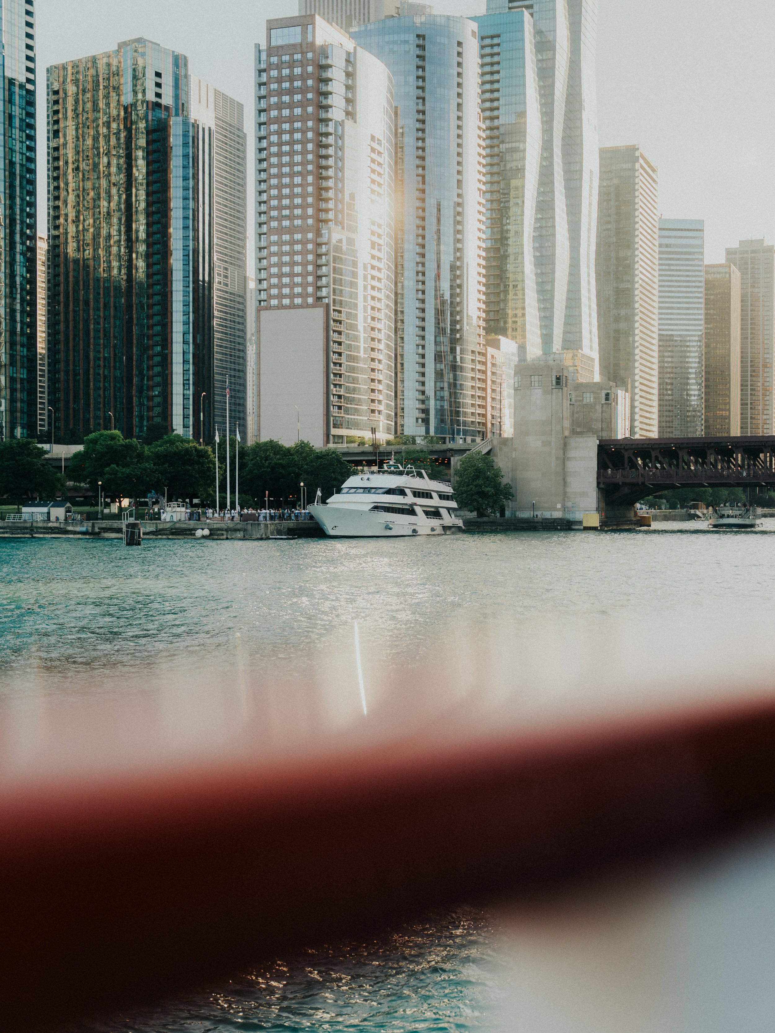Cityscape with tall modern skyscrapers along a river, featuring a white yacht docked along the waterfront and a bridge crossing over the river in the foreground.