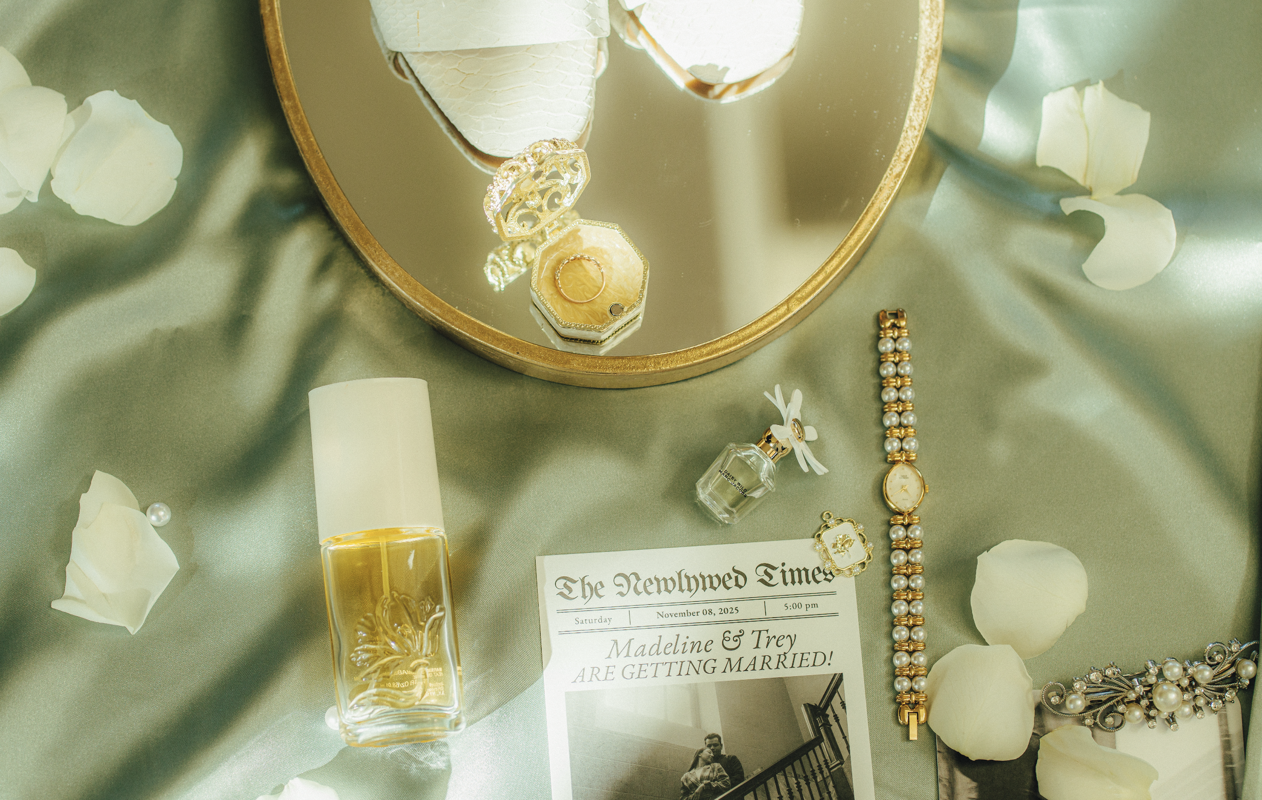 A mirror reflecting jewelry including a gold ring and a pendant, a perfume bottle with a ribbon, a pearl watch, pearl earrings, a floral headpiece, and white flower petals on a green satin cloth, with a wedding announcement pointing out that Madeline and Trey are getting married on November 8, 2025.