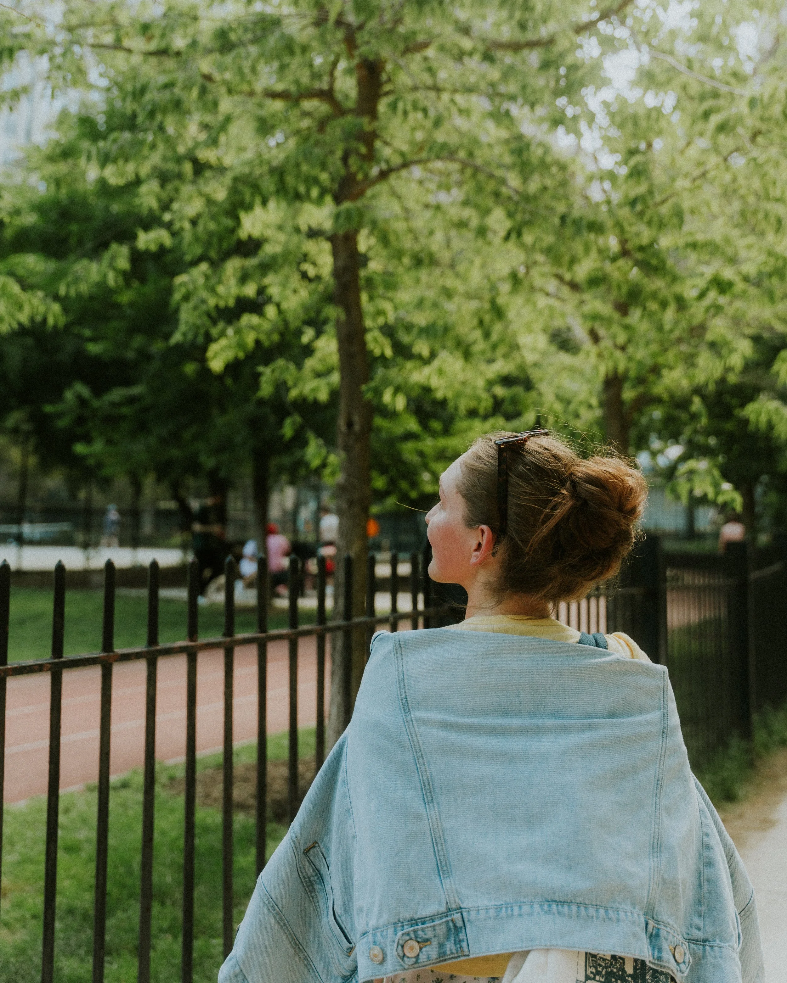 Woman with her hair in a bun, wearing a denim jacket, sitting outdoors near a black fence with green trees in the background.