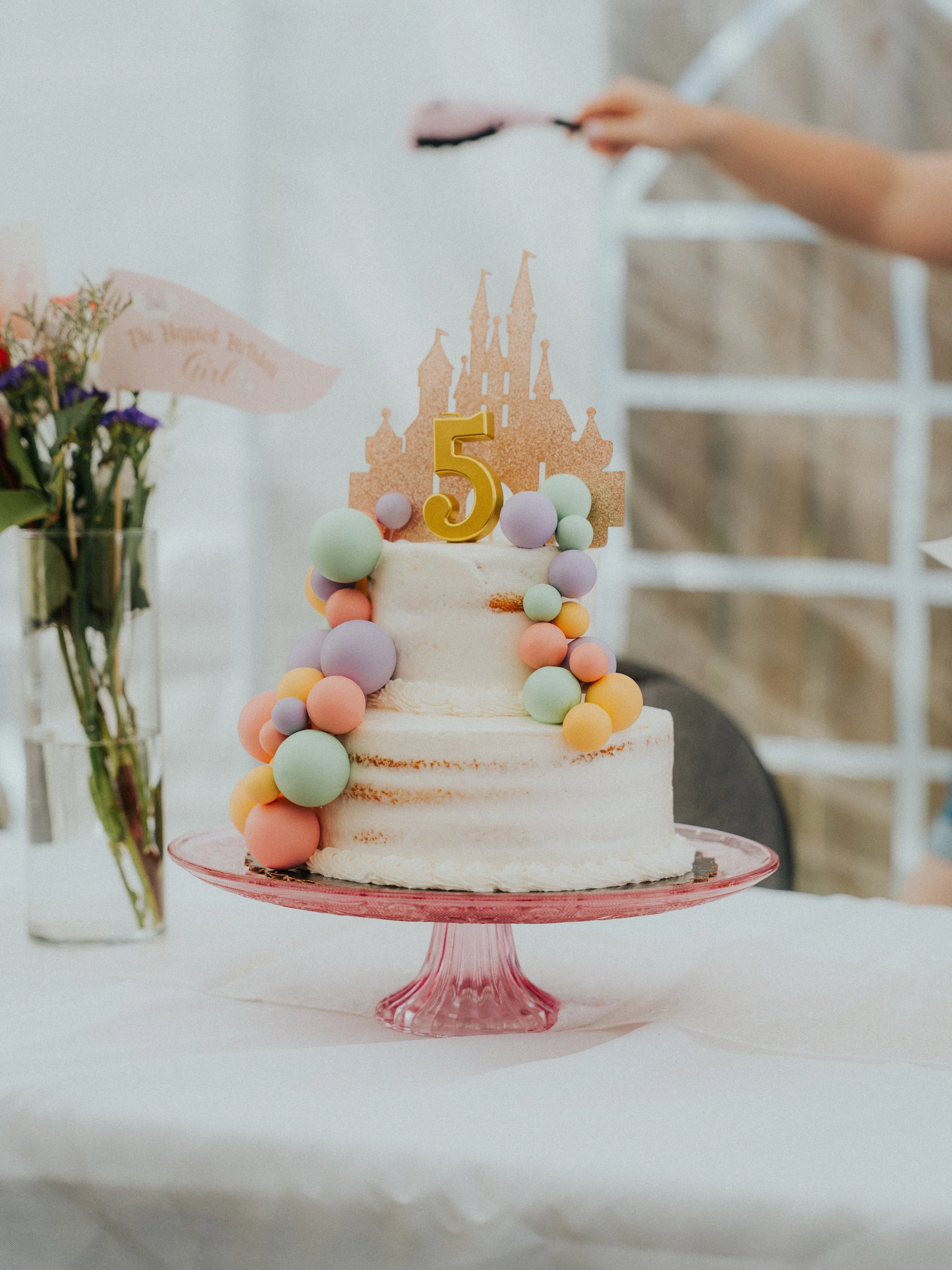 A two-tiered white birthday cake decorated with pastel-colored spheres and a gold number 5 topper, with a pink castle topper on top, placed on a pink glass stand, on a white table with a floral arrangement nearby.