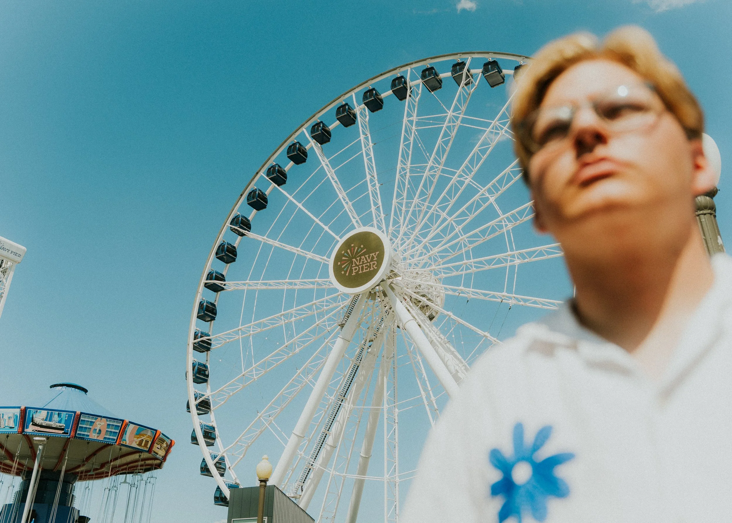 A large Ferris wheel with a sign that says 'Navy Pier,' against a clear blue sky, with a slightly blurred person in the foreground.