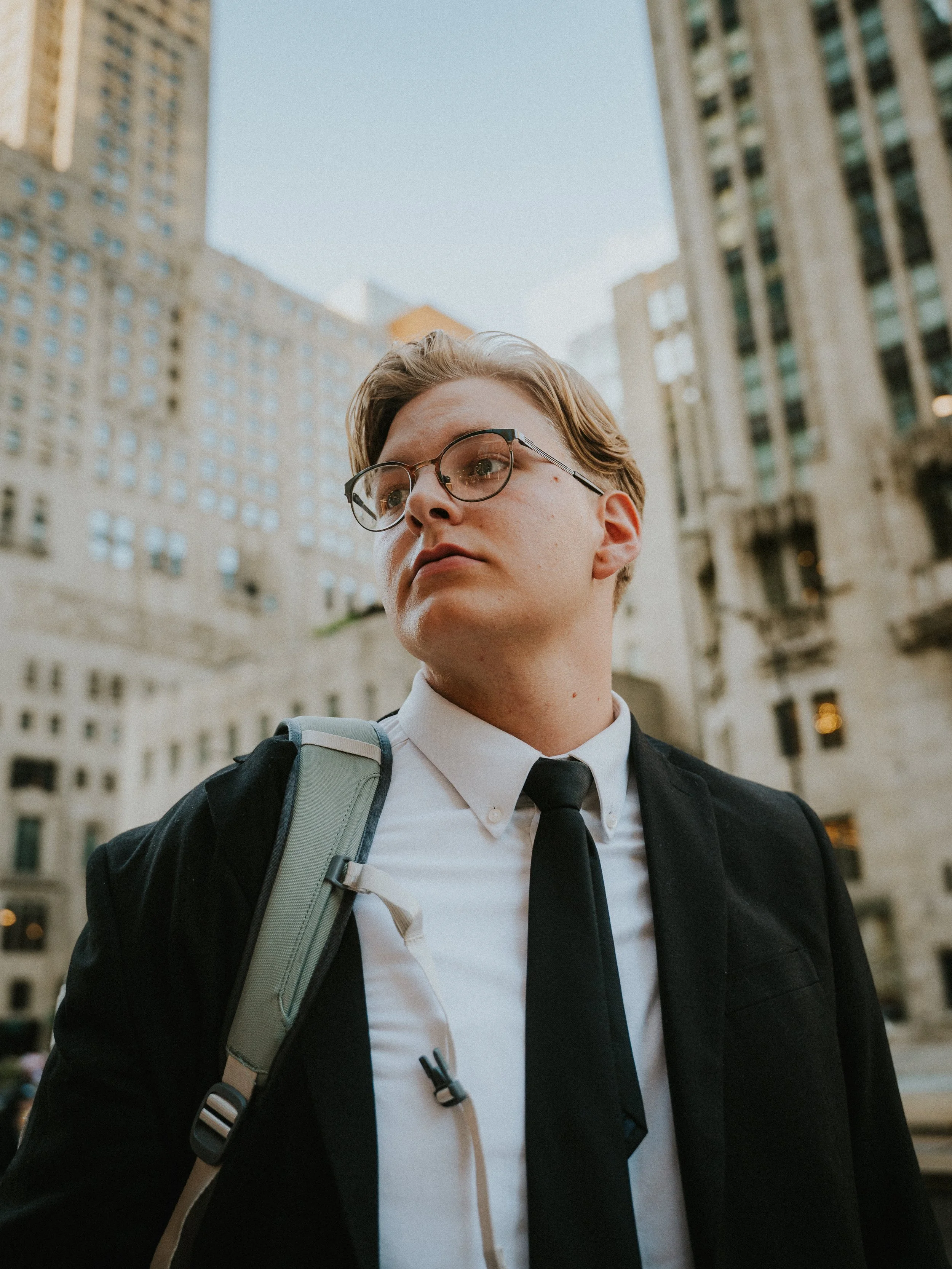 Young man with glasses wearing a white shirt and black tie, carrying a backpack, standing in front of tall city buildings.