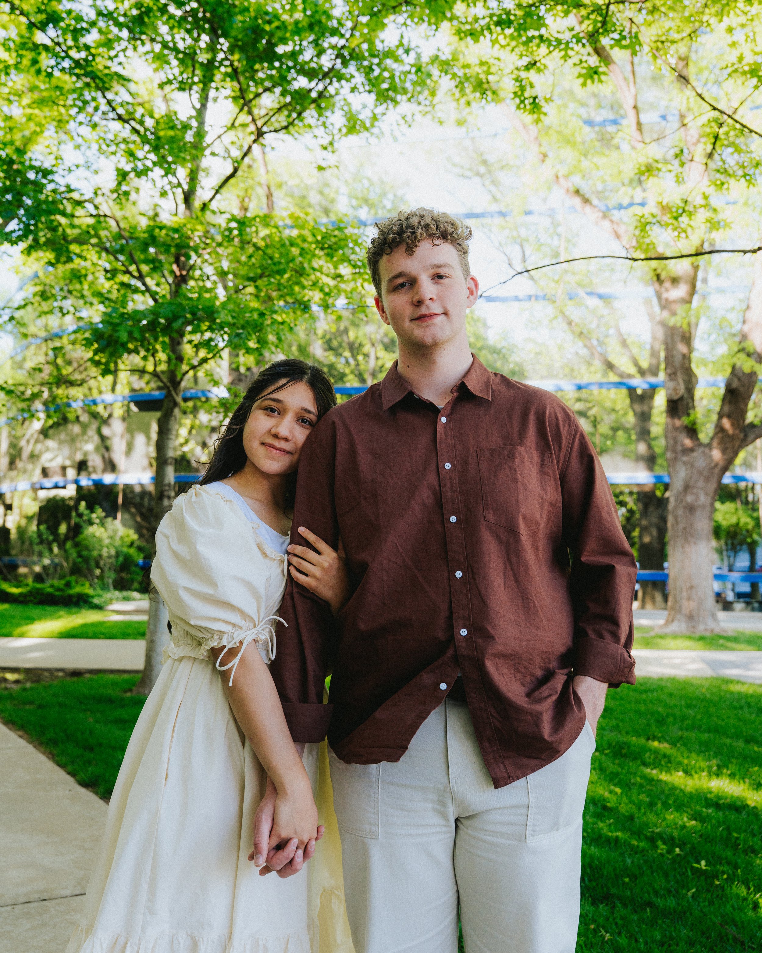 A young couple standing close together outdoors, surrounded by green trees and grass on a sunny day.