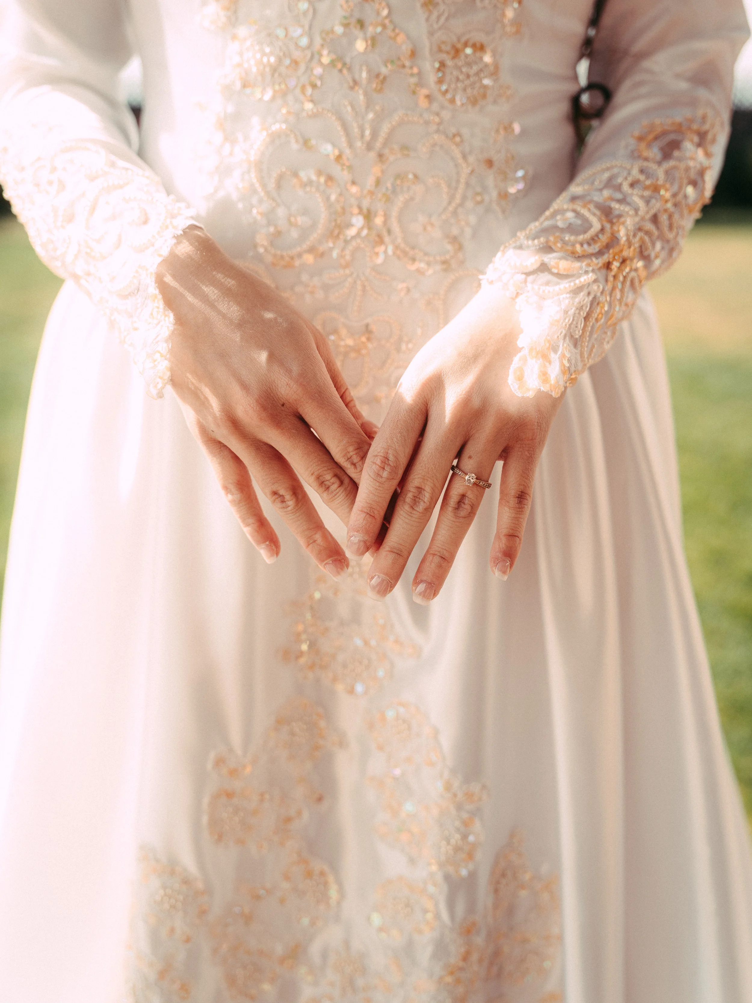 Close-up of a bride's hands displaying engagement and wedding rings, wearing a white wedding dress with gold embroidery and lace sleeves.