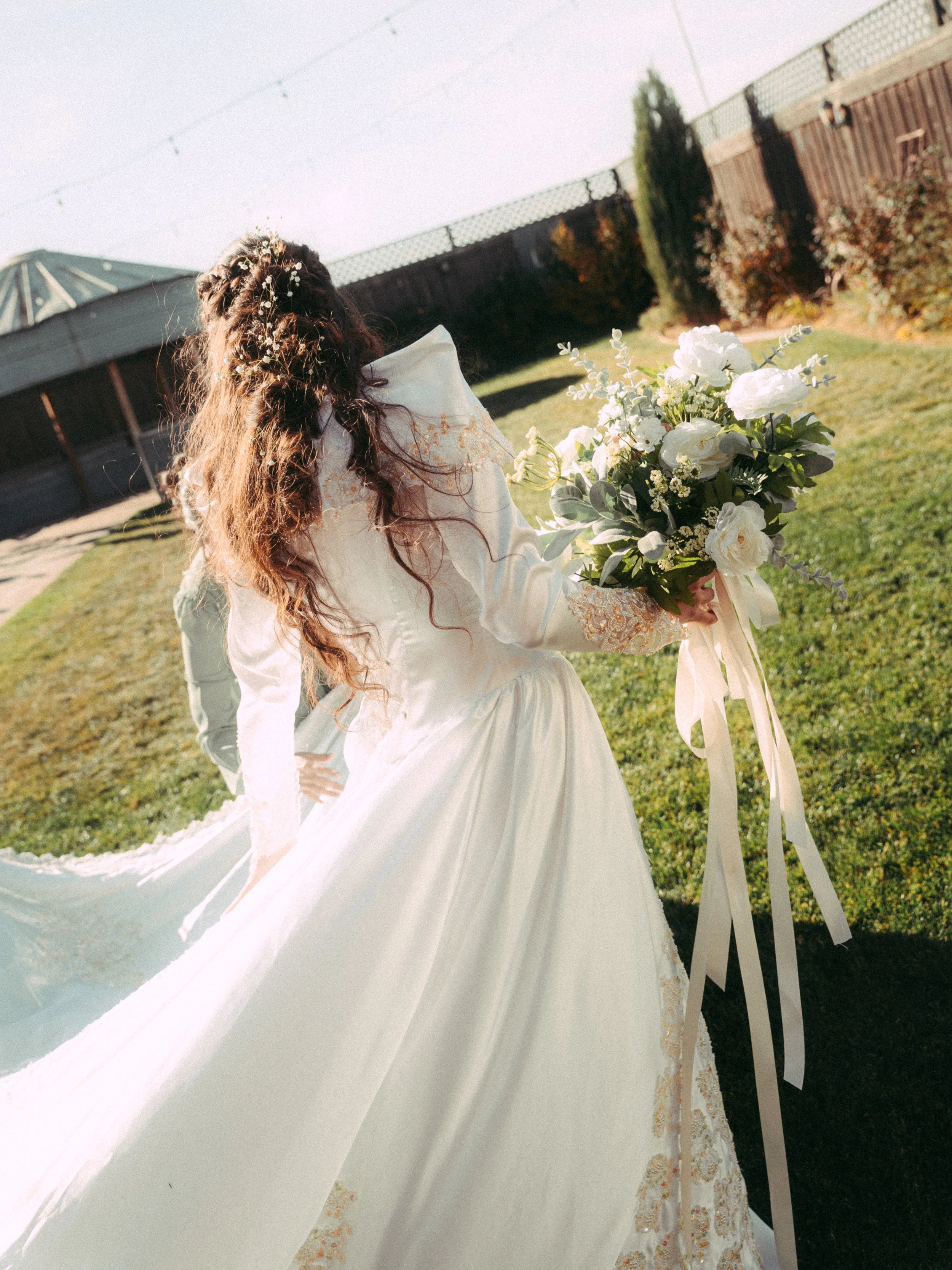 A bride in a white wedding dress holding a bouquet of white flowers outdoors in a backyard with green grass, a fence, and trees.