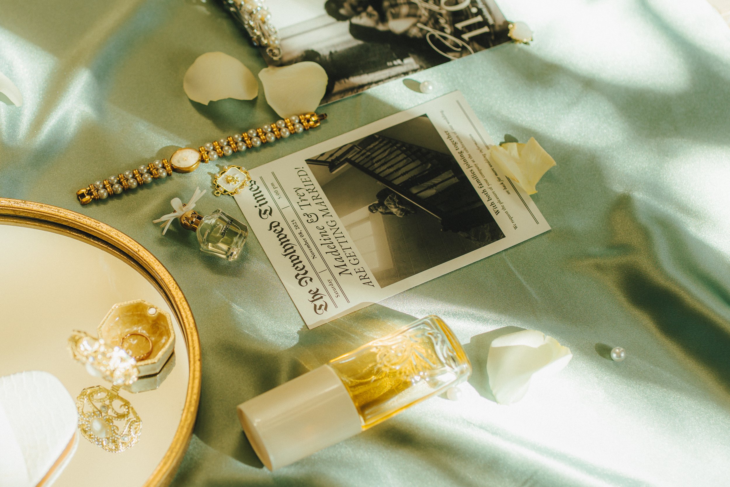 A flat lay of a wedding or engagement theme with jewelry, perfume, flower petals, and a newspaper on a light green satin fabric.