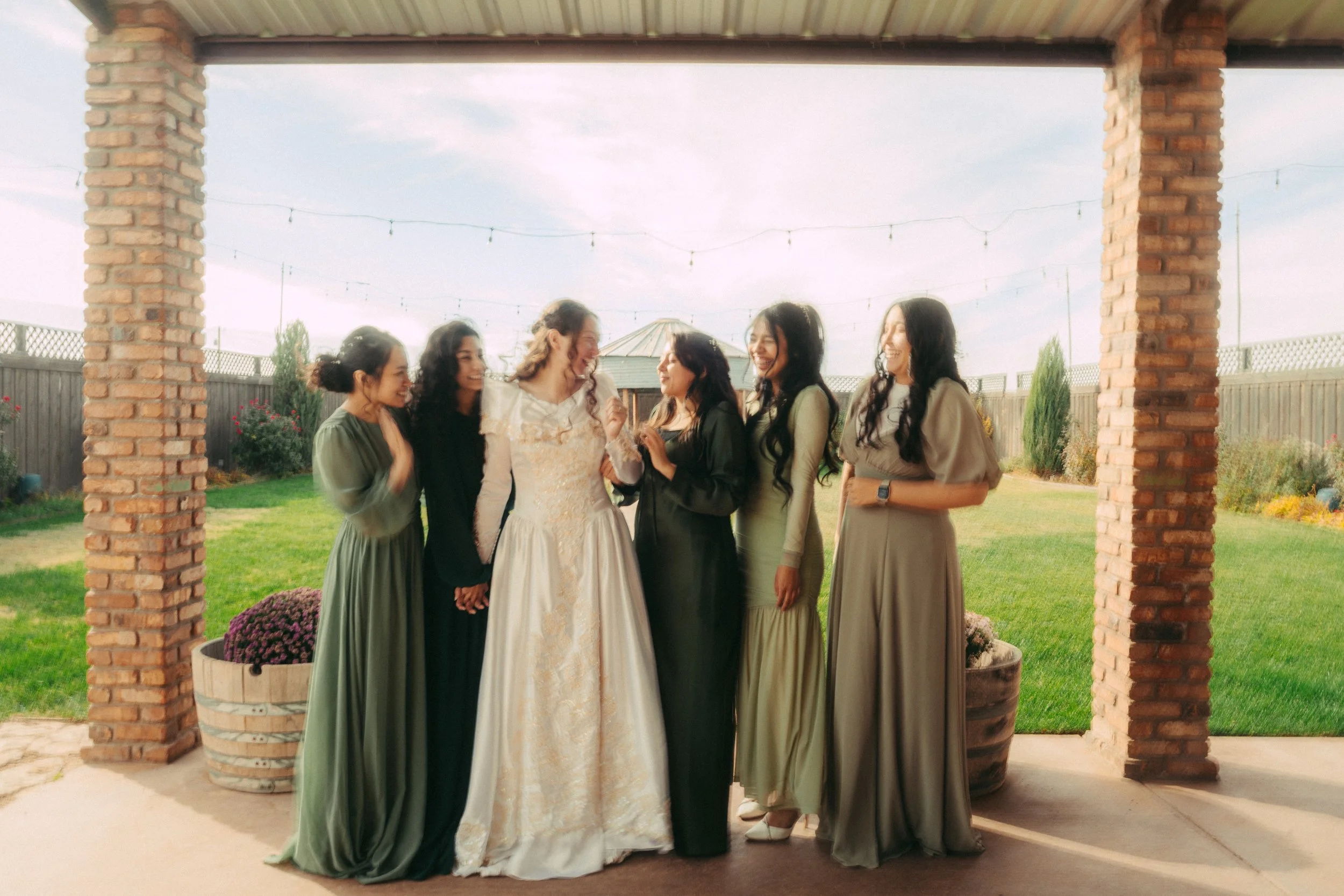 A group of six women, including one in a white wedding dress, standing outdoors on a patio under a covered structure, smiling and laughing together during a celebration, with a grassy yard, potted plants, and string lights in the background.