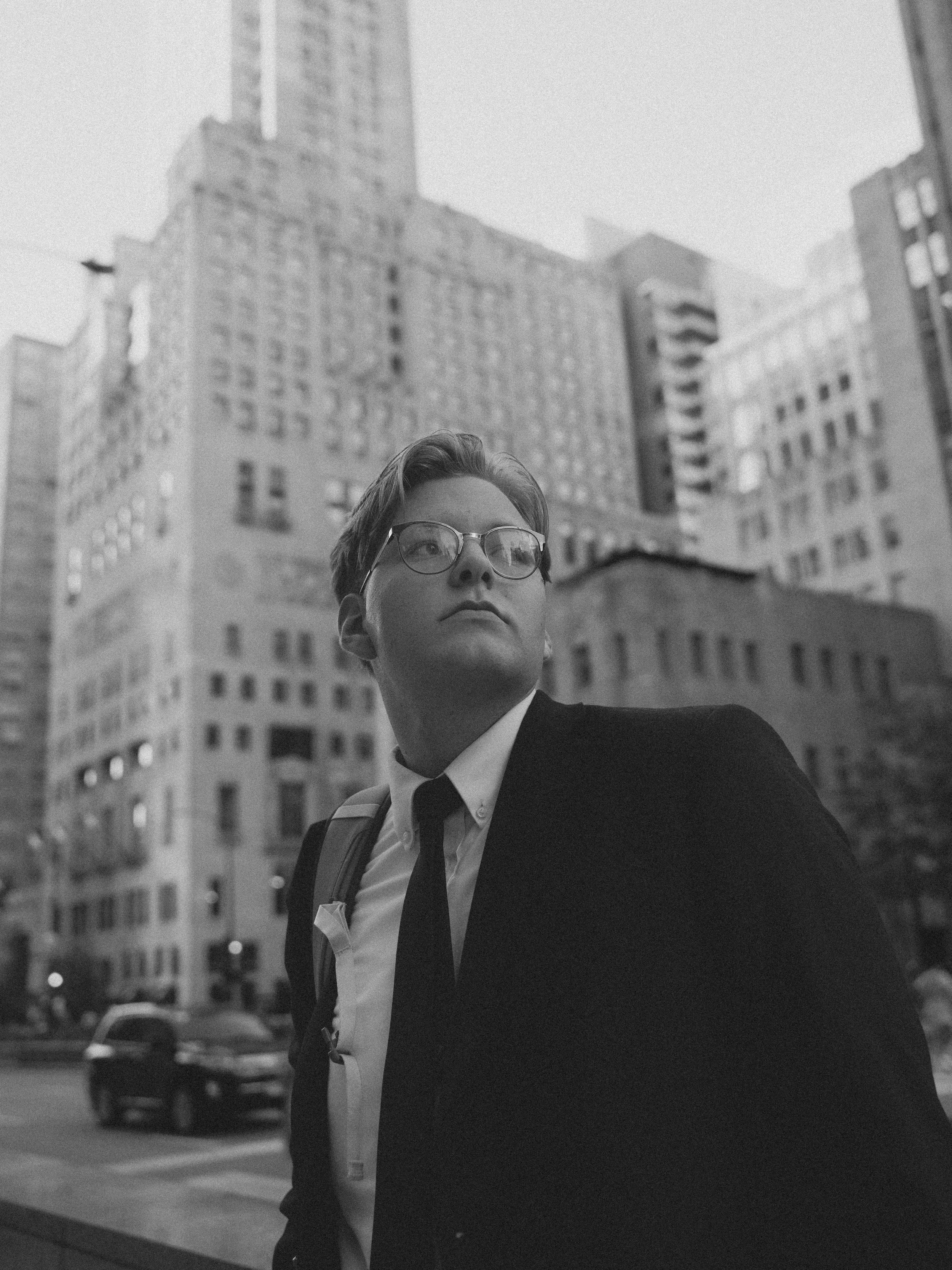 A young man in a suit and glasses looking upward in an urban city with tall buildings in the background.