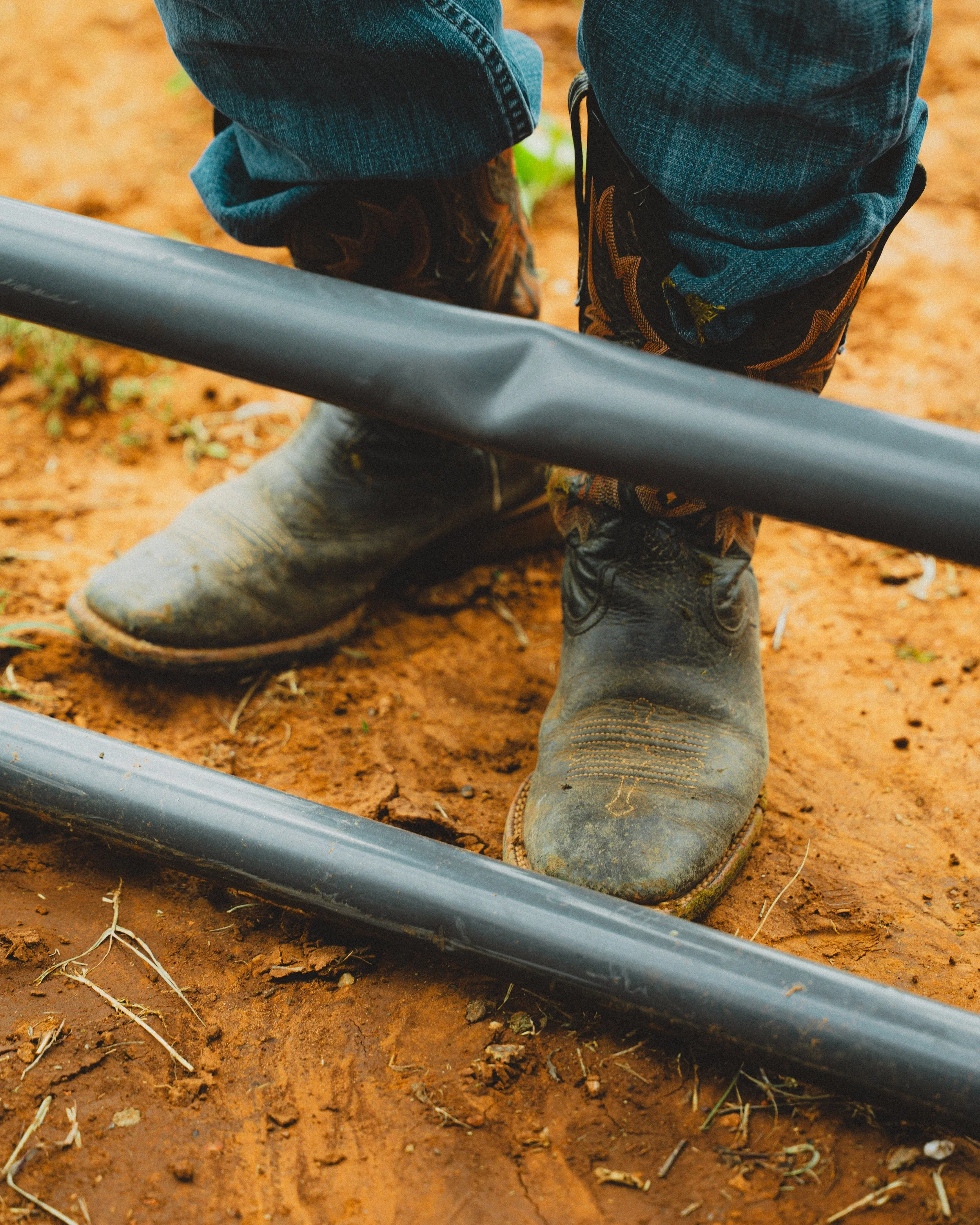 Close-up of a person's feet in muddy cowboy boots, standing on red dirt, with denim jeans, near metal pipes on the ground.