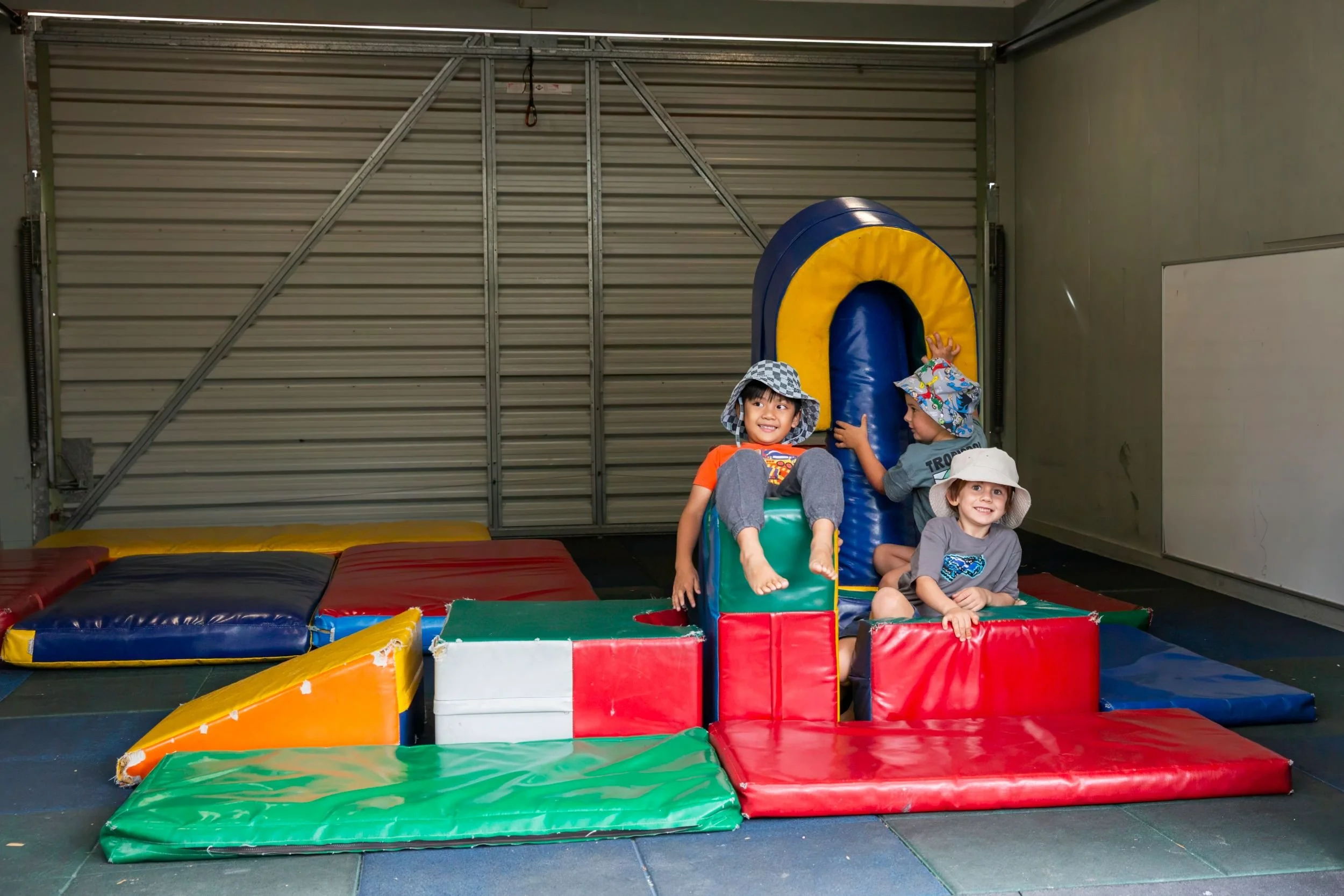 Three young children playing on colorful indoor bounce house mats and inflatable structures, with a garage door in the background.