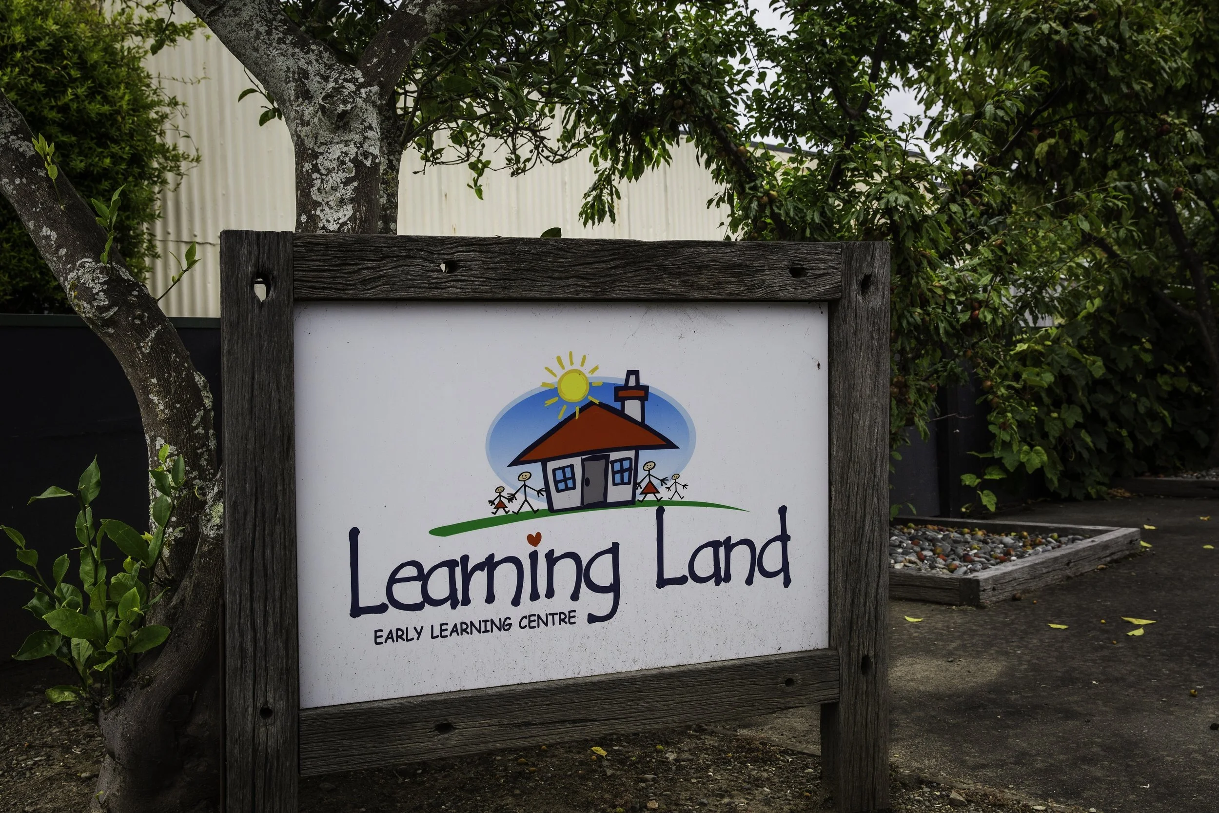 Sign for Learning Land Early Learning Centre featuring a house with children and sun illustration on a whiteboard framed with weathered wood, outdoors with trees and shrubs in background.