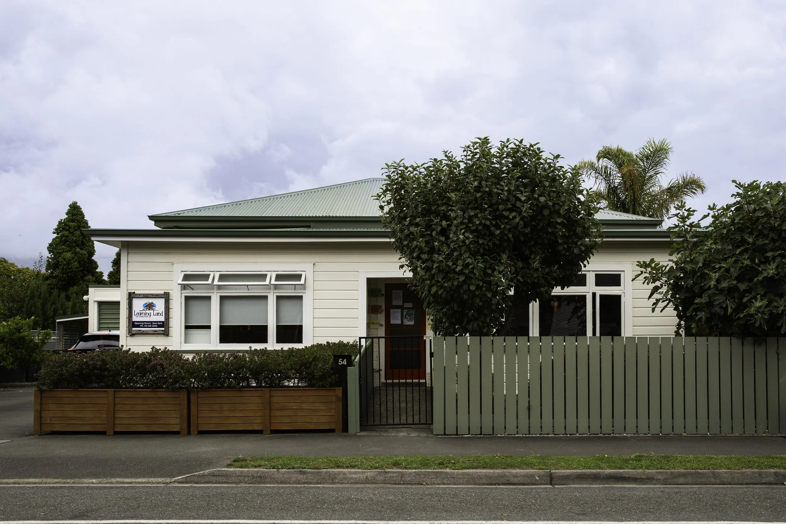 Front view of Learning Land with a green metal roof, large windows, a small red framed front door. An apple tree and shrubs in the front, a wooden fence, with a grassed curb. The sky is cloudy.