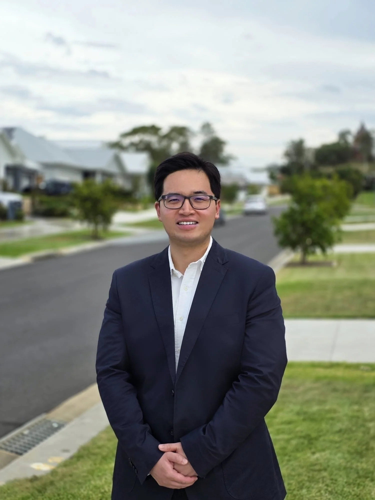 A man in a black suit and white shirt standing outdoors on a residential street with houses, trees, and a cloudy sky in the background.