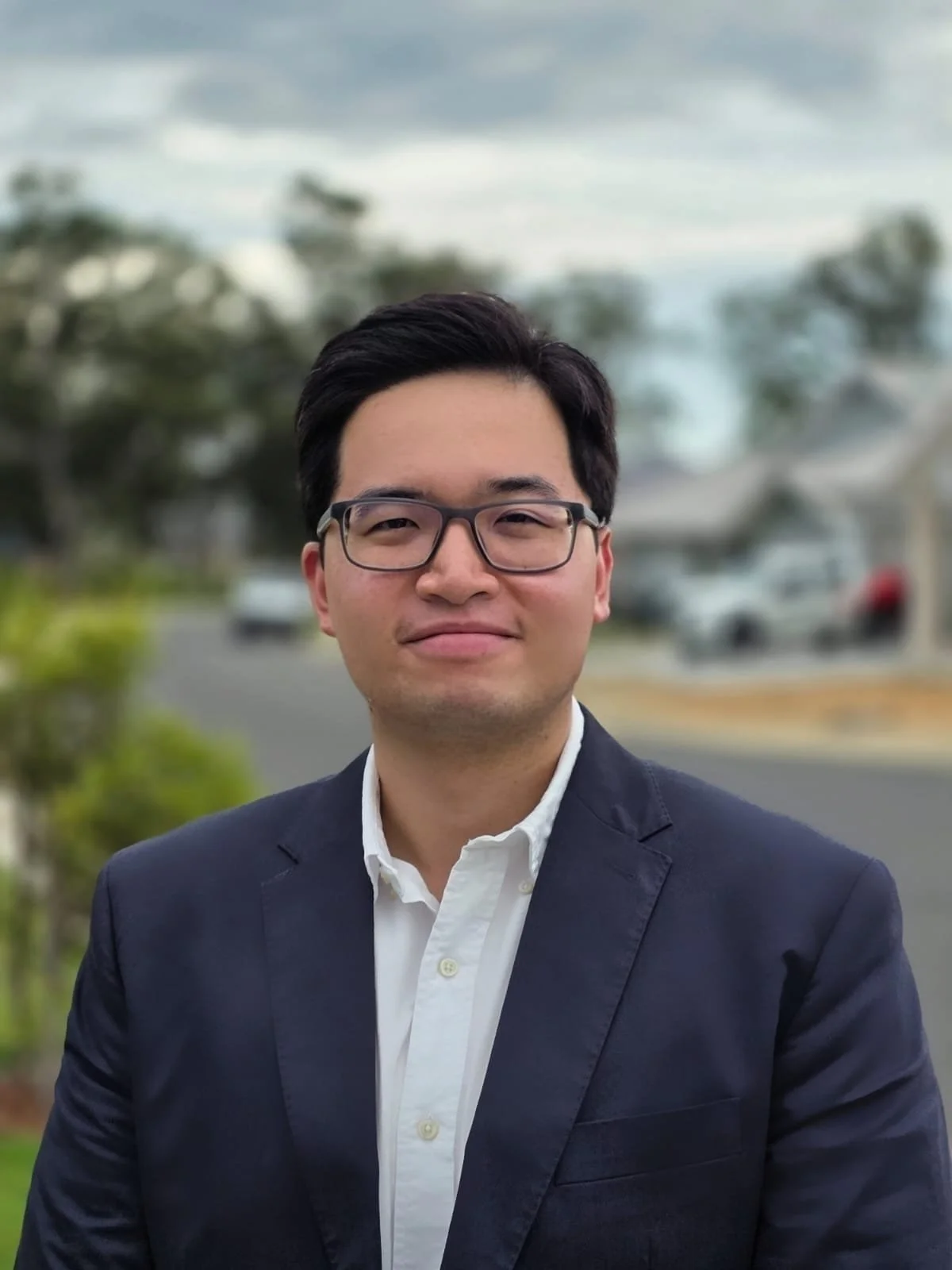 A man in a dark blazer and white shirt standing outdoors, with a blurred background of trees, houses, and parked cars.