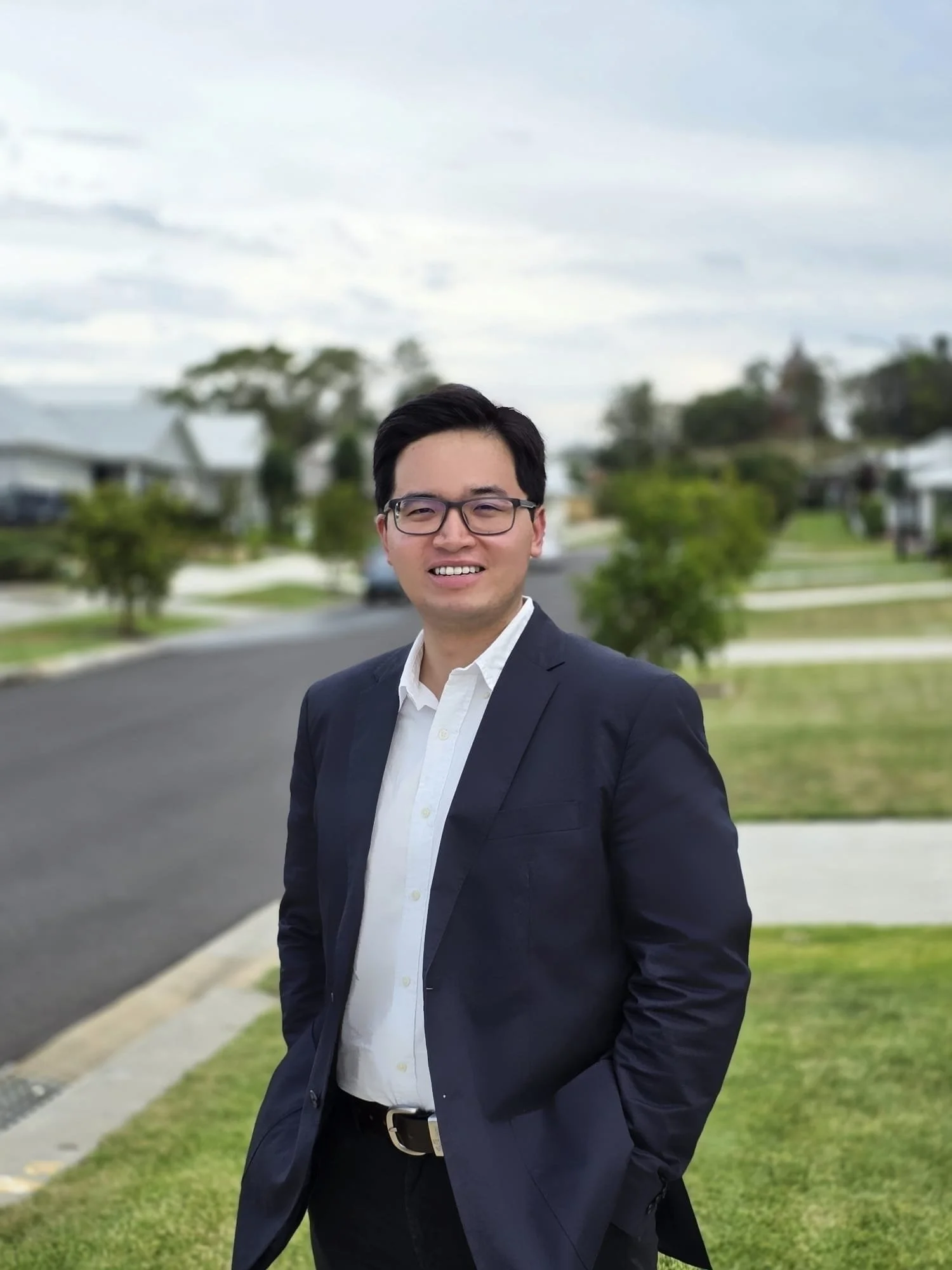 A young man with dark hair, glasses, wearing a white shirt and dark suit, standing on a suburban sidewalk with a grassy yard and trees in the background.