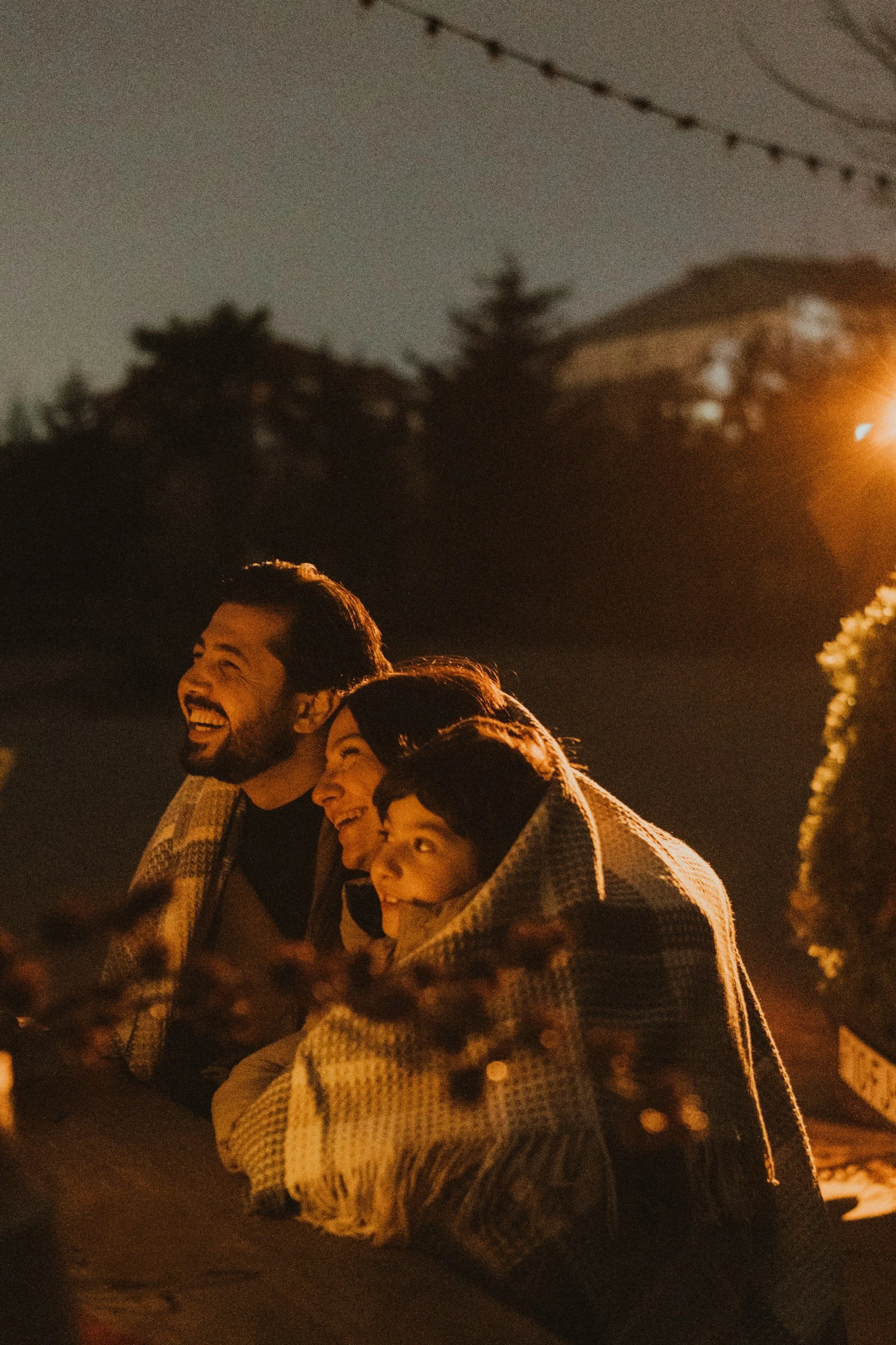 Family of four smiling and sitting outdoors at night around a campfire, wrapped in blankets, with trees and a house in the background.