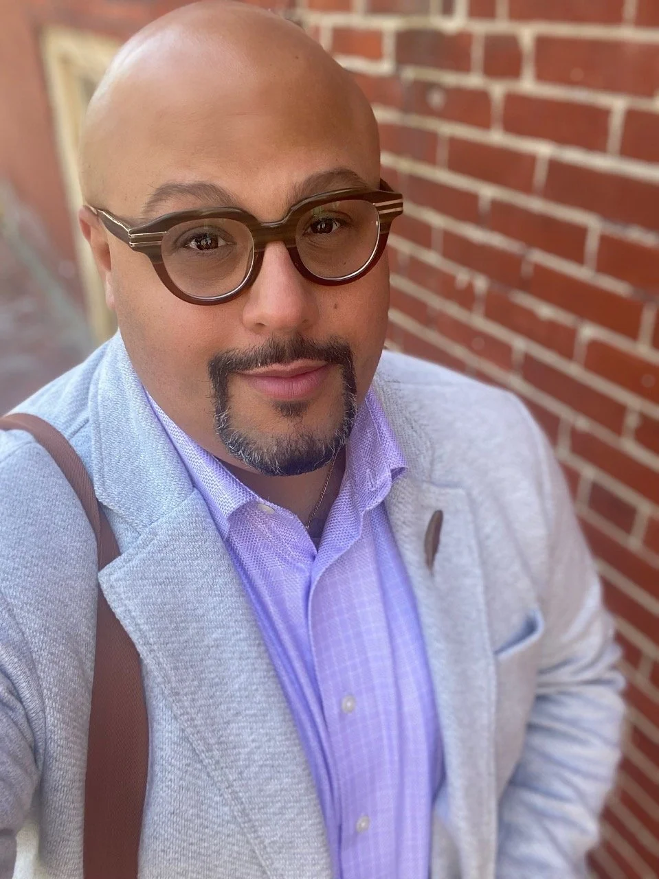 A man with a shaved head, glasses, and a goatee smiling for a selfie outside, with a brick wall in the background.