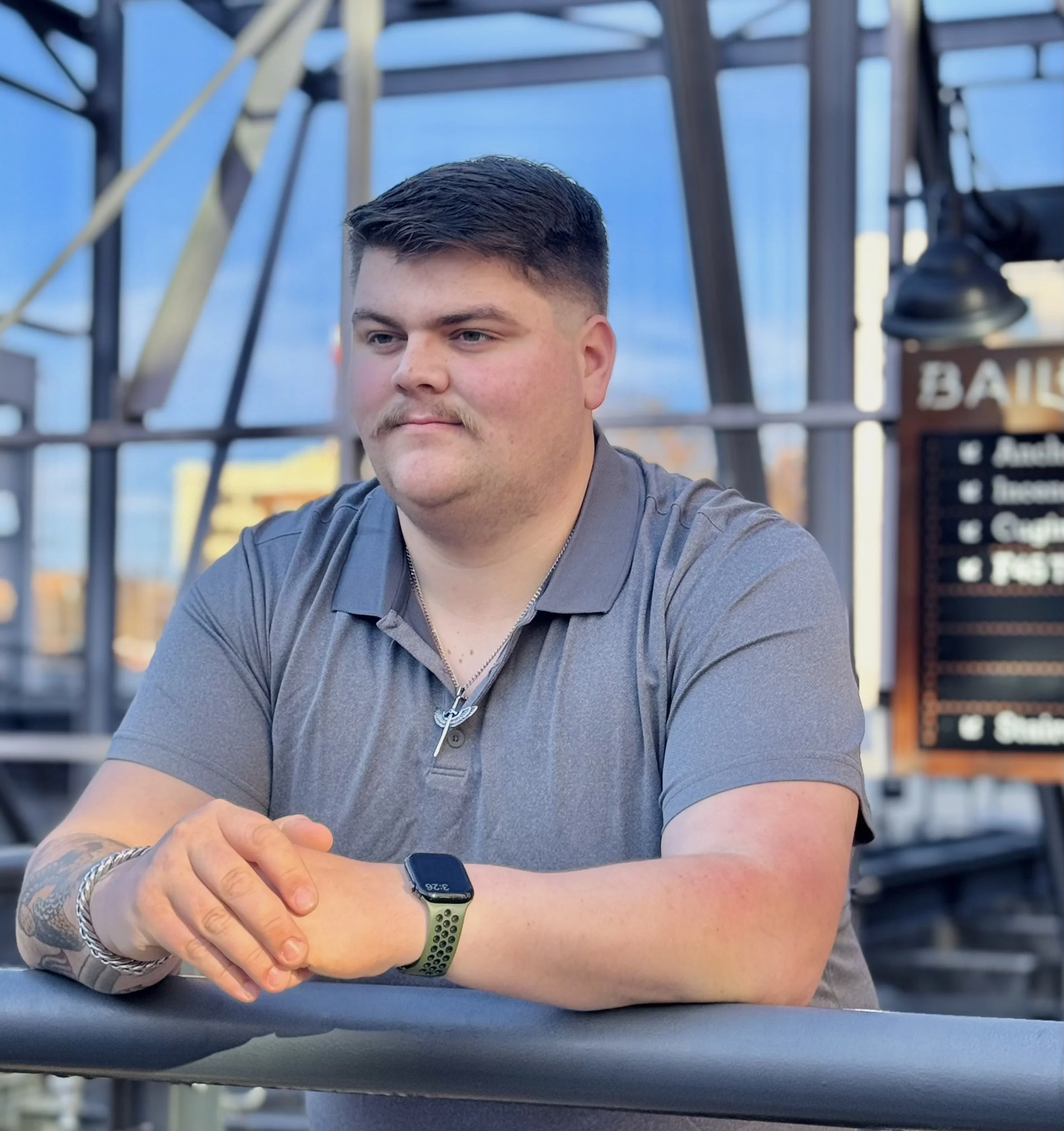 A young man with short dark hair, a mustache, and light skin sits at a table outside, wearing a gray polo shirt, a silver chain, a silver bracelet, and a smartwatch. He appears to be in a modern outdoor space with metal framework and a sign in the background.
