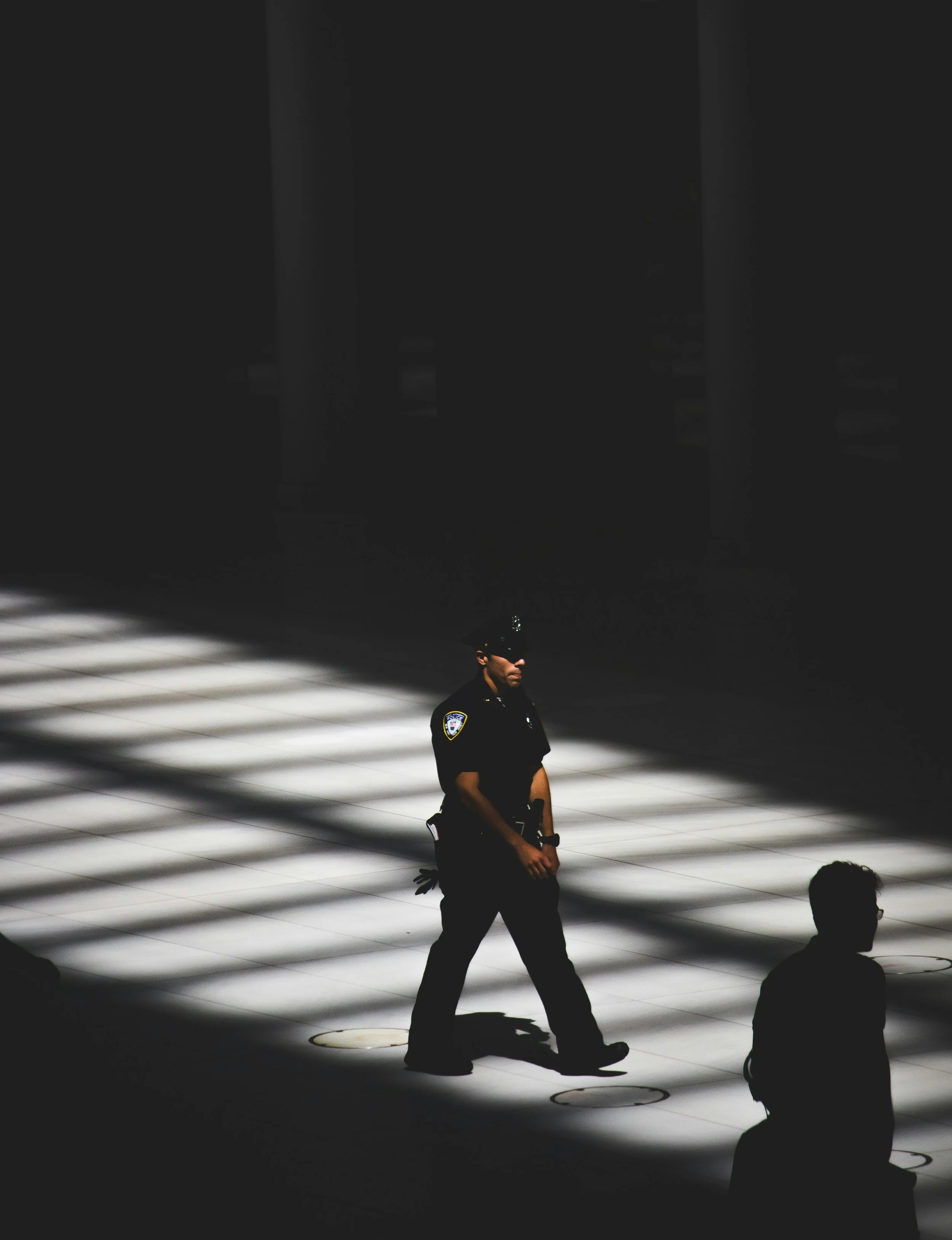 A police officer standing in a brightly lit area, with shadows and silhouette figures around him in darkness.