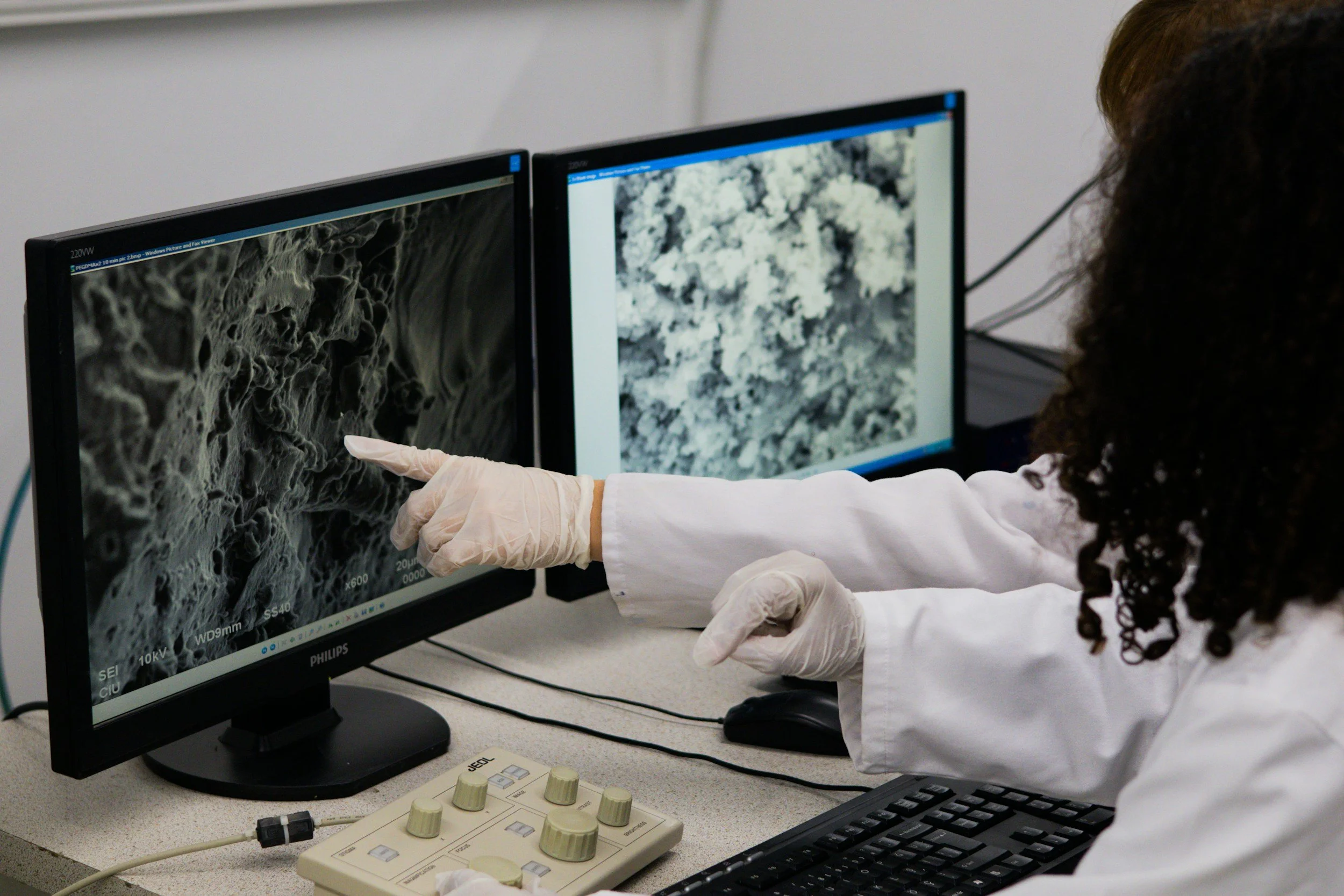 Researcher pointing at electron microscope images on two computer monitors in a laboratory.