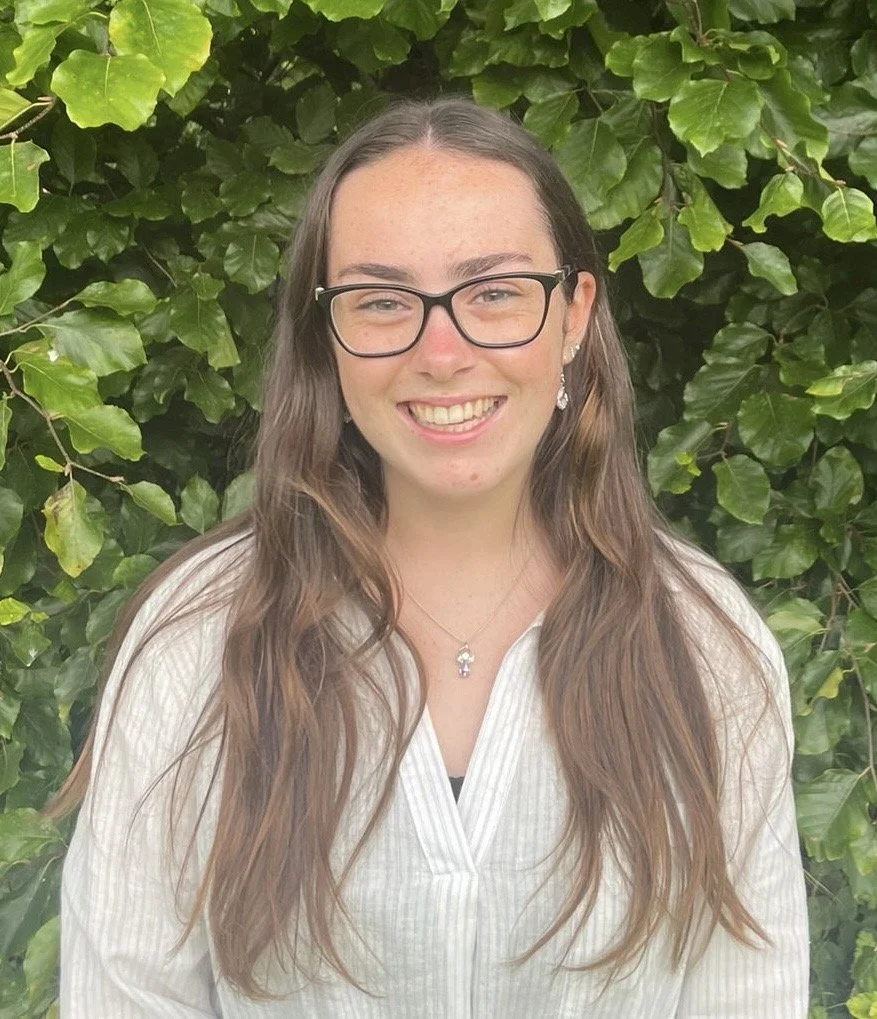 A young woman with long brown hair wearing glasses, earrings, and a necklace, standing outdoors in front of green leafy foliage, smiling at the camera.