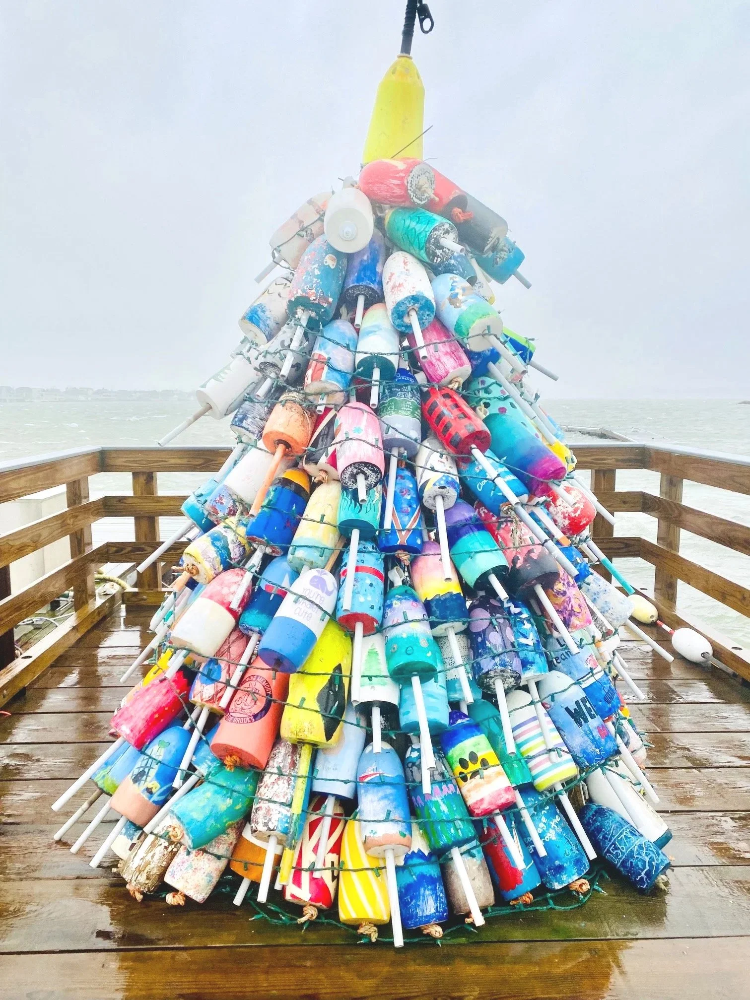 A large, colorful sculpture made of painted buoys stacked in a conical shape on a wooden pier with a foggy water background.