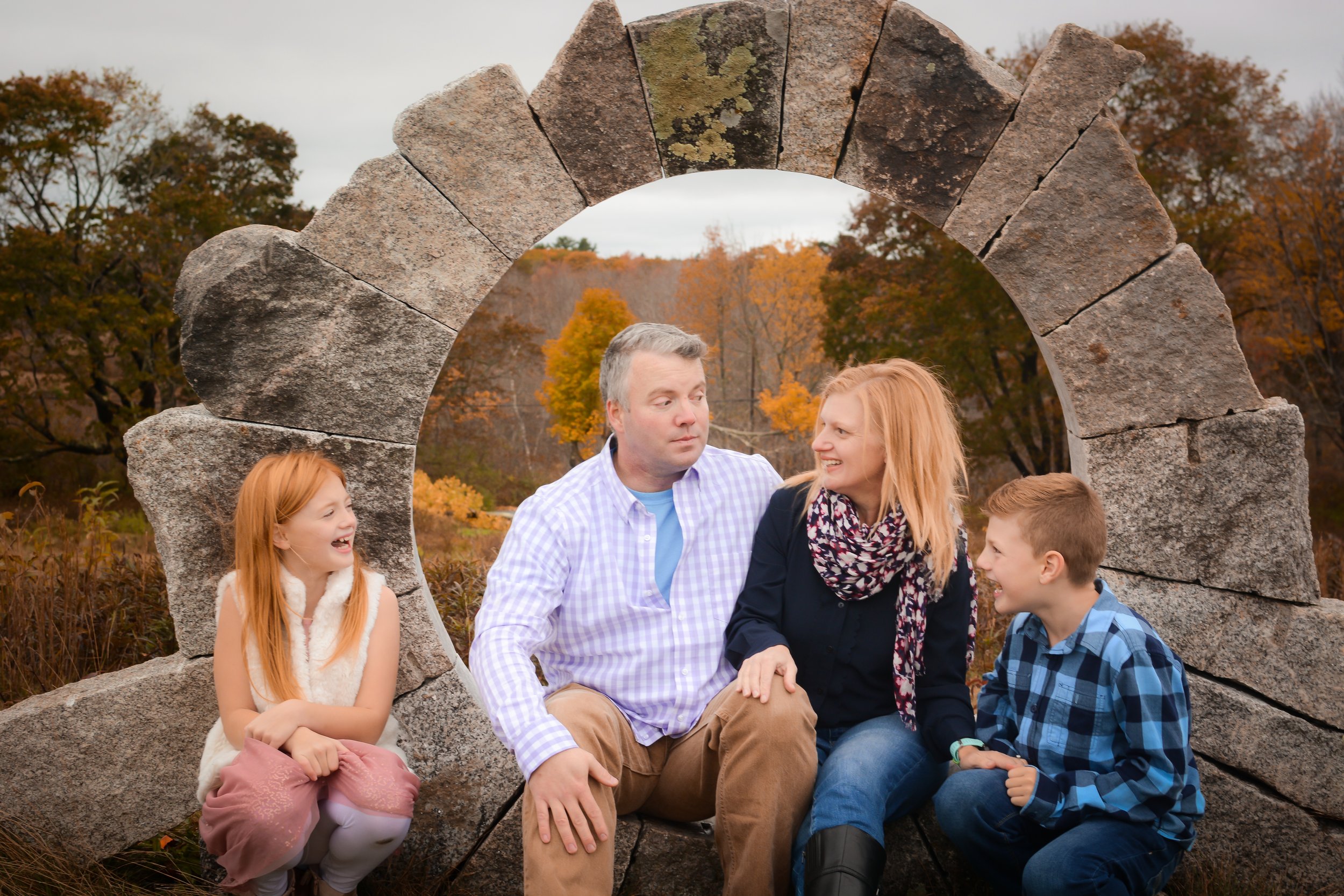 A family of four sitting on a stone bench under an arch in a park during fall, with colorful autumn trees in the background. The parents are talking and smiling, while the children are laughing.