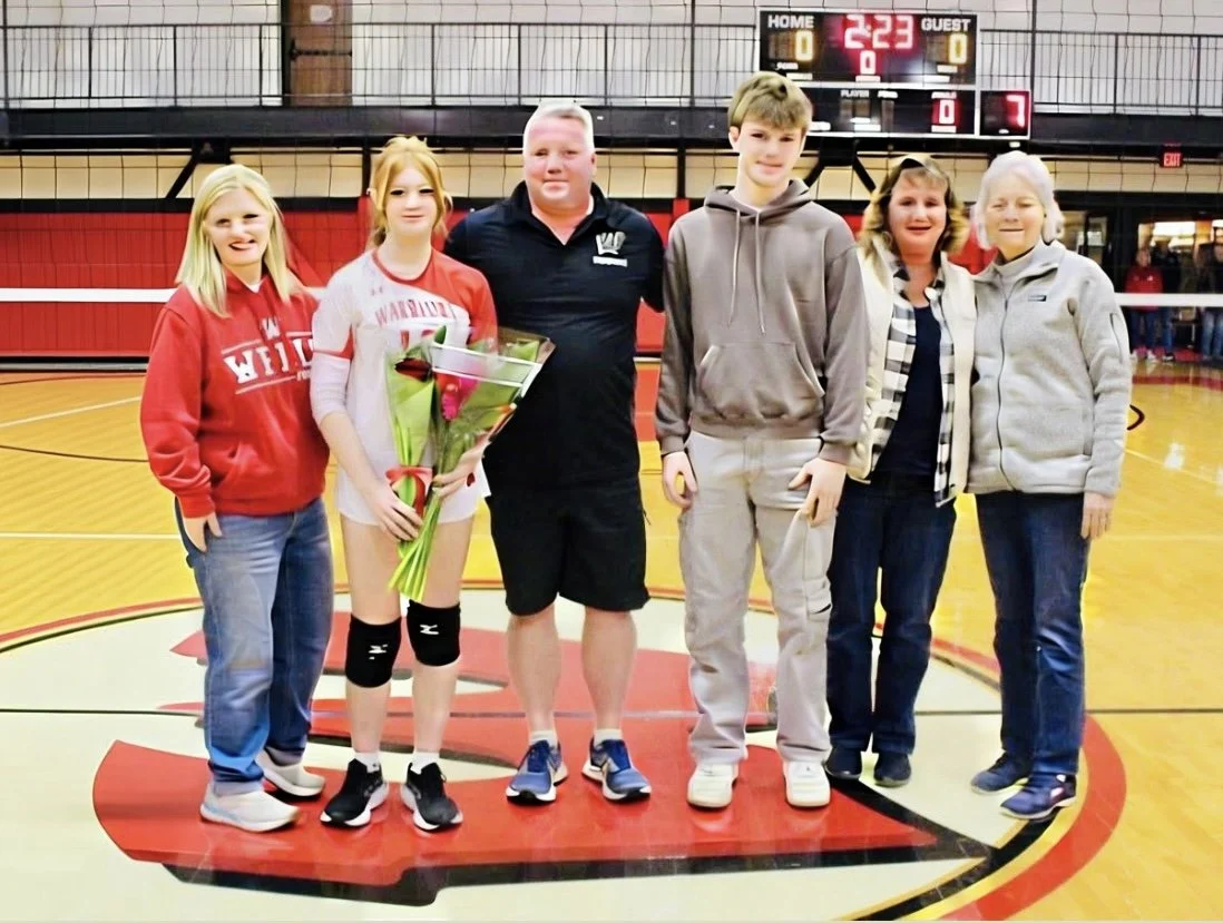 Group of six people, including a girl with a bouquet of flowers, standing on a gymnasium basketball court with a scoreboard in the background.