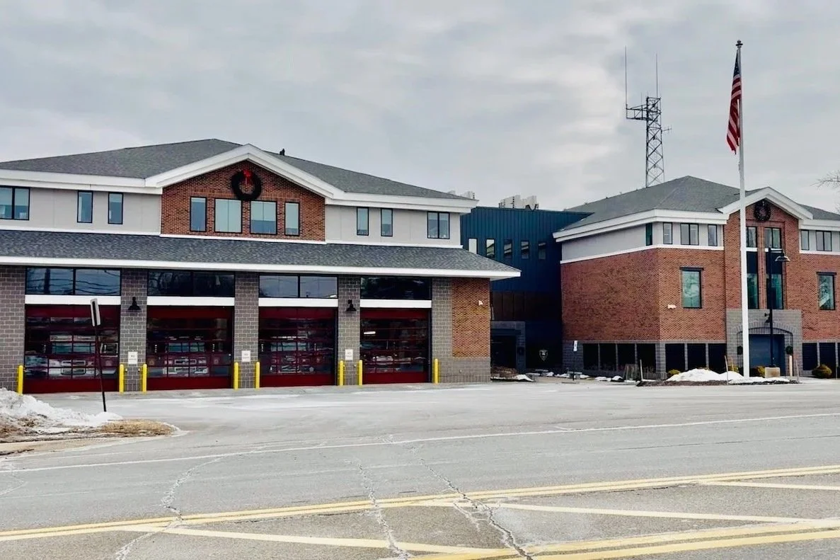 Fire station with three garage bays, brick and white siding, and American flag in front, winter scene with snow on ground.