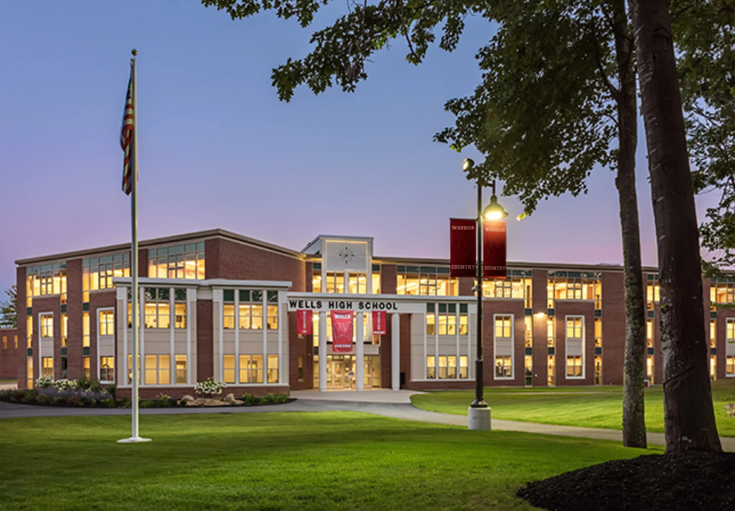 Exterior view of Wells High School at dusk, showing the building's brick facade, lit windows, American flag, and surrounding trees and grass.