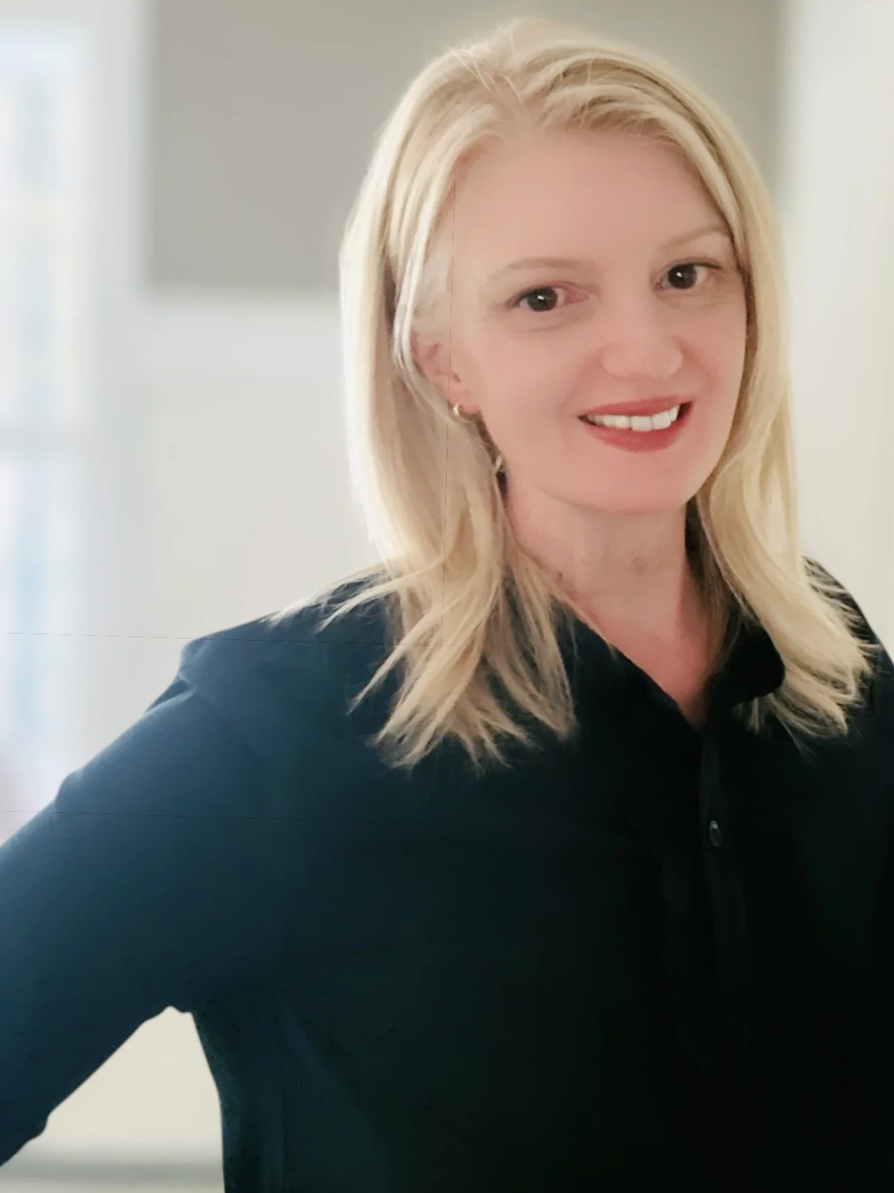A woman with blonde hair smiling, wearing a black top, indoors with a blurred background.