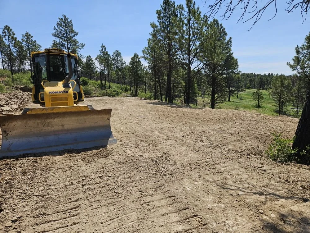 A yellow Komatsu bulldozer working on a dirt road surrounded by trees and clear blue sky.