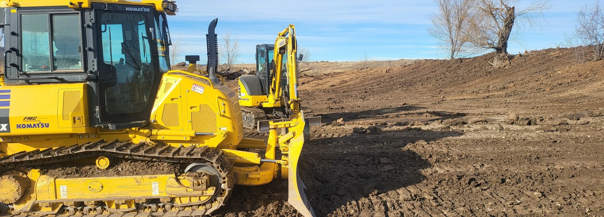 A yellow Komatsu bulldozer working on a dirt field with a clear blue sky and a few trees in the background.