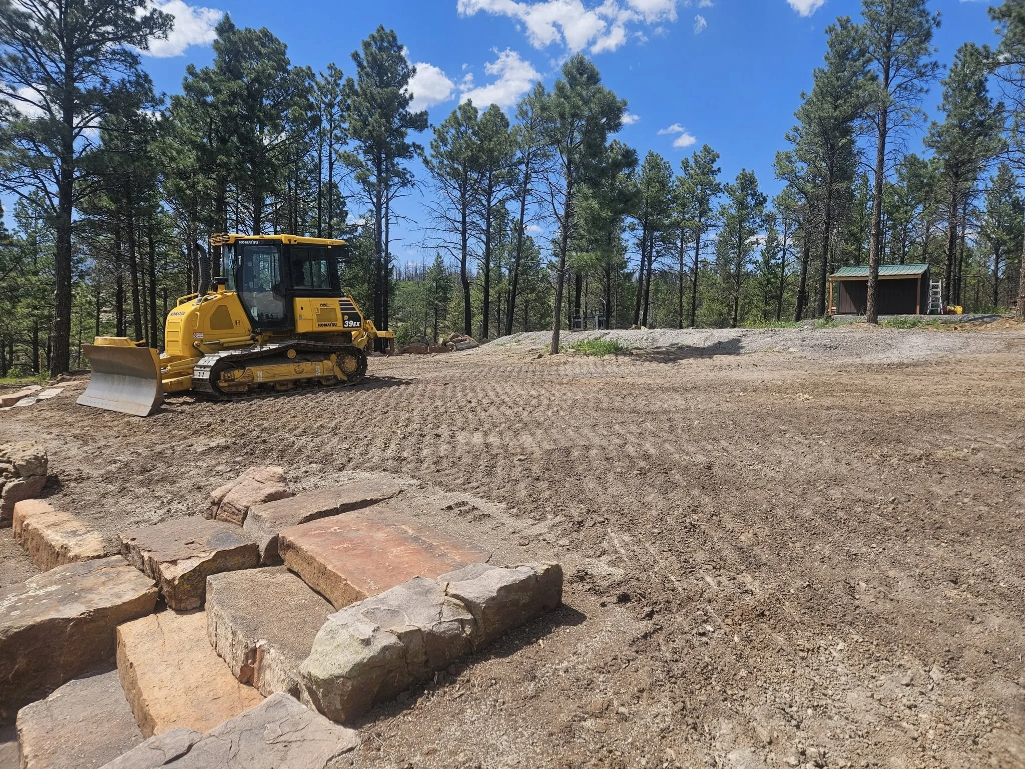 A yellow bulldozer parked on a dirt site with a forest of tall pine trees and a small building in the background, under a blue sky with some scattered clouds.