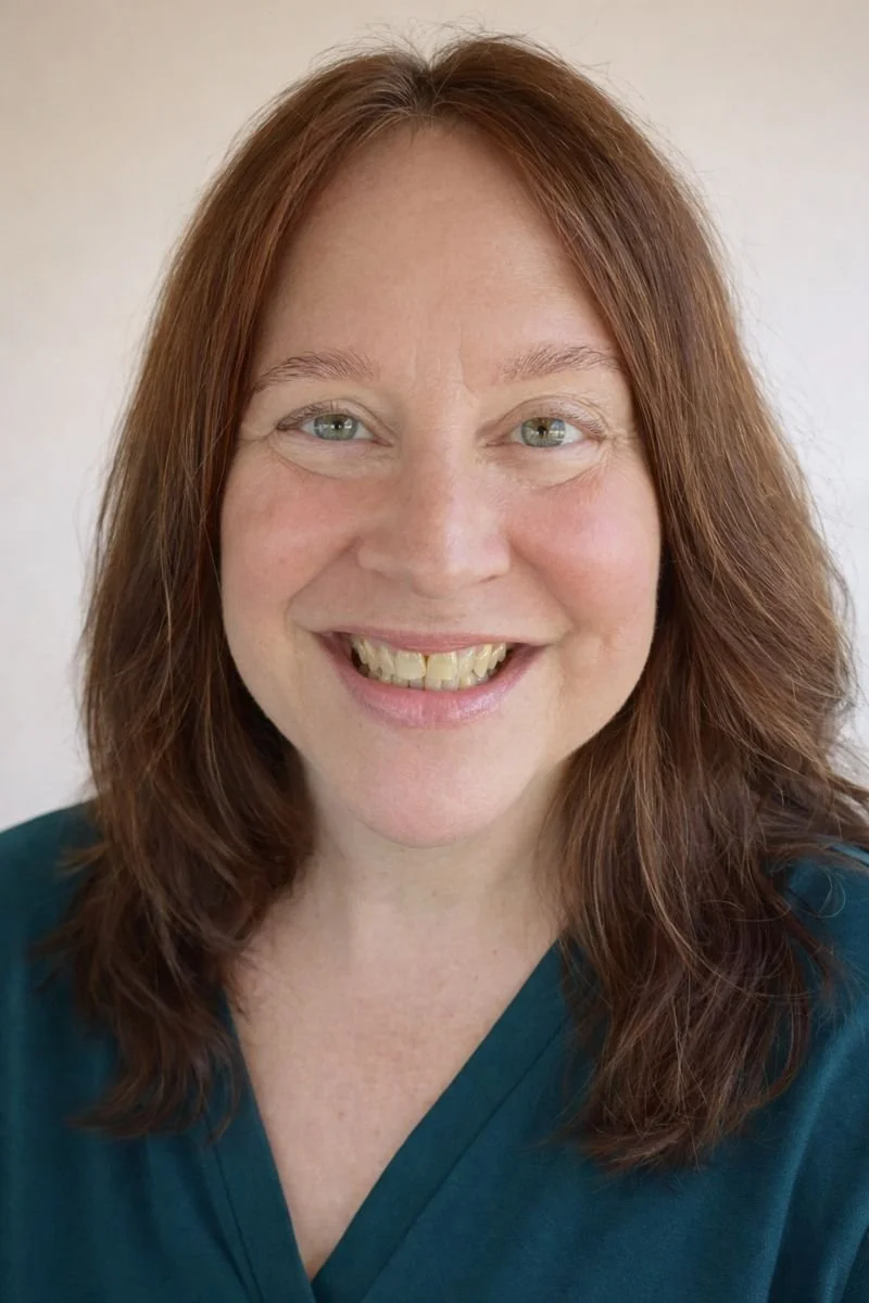 Close-up of a woman with auburn hair, hazel eyes, and a big smile, wearing a dark teal top.