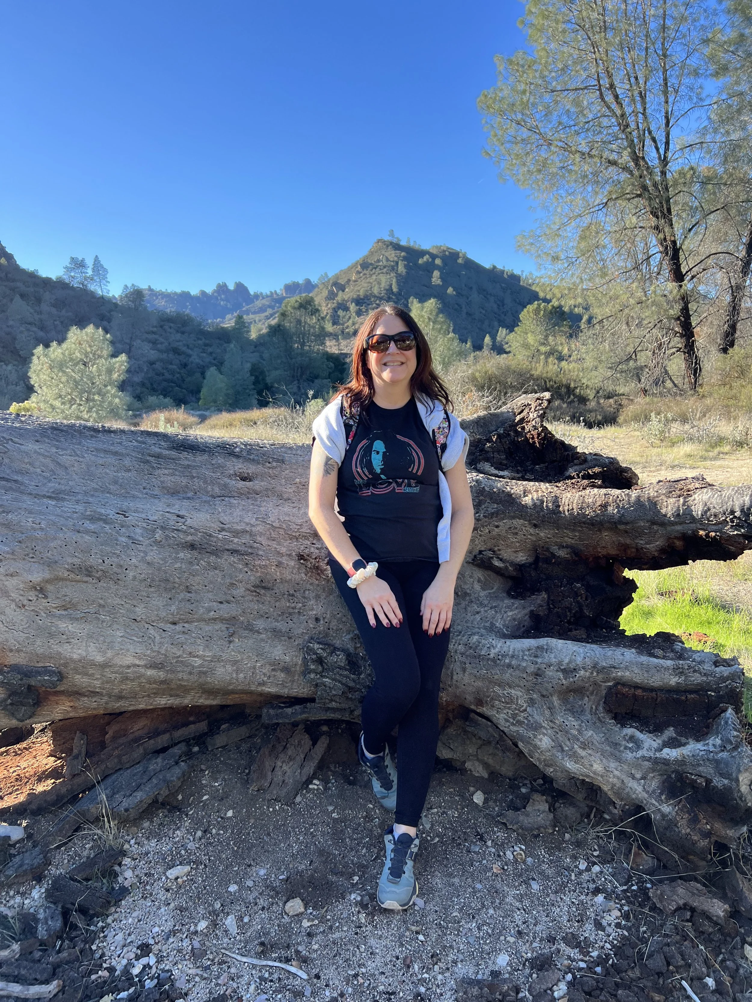 A woman with sunglasses and a black graphic t-shirt sitting on a fallen tree in a mountainous outdoor setting with green trees and a clear blue sky.