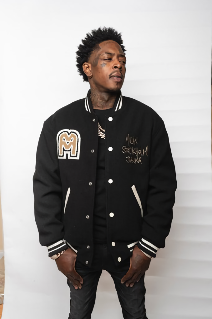 A young man with tattoos and curly hair wearing a black varsity jacket with patches and text, standing against a white background.