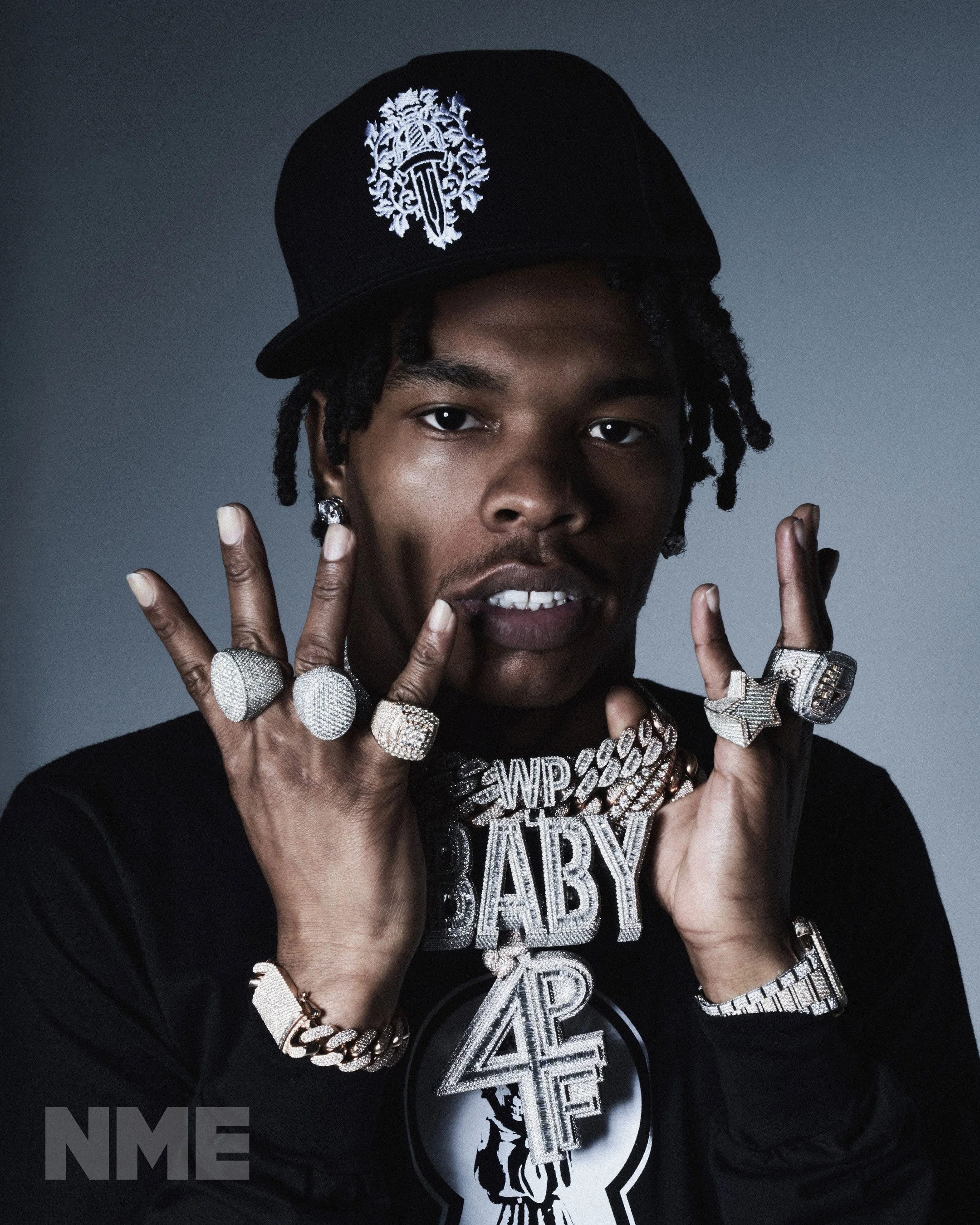A young man wearing a black baseball cap with white embroidery, multiple jewelry including rings, bracelets, and necklaces, and a black shirt with graphic design, posing with his hands near his face against a dark gray background.