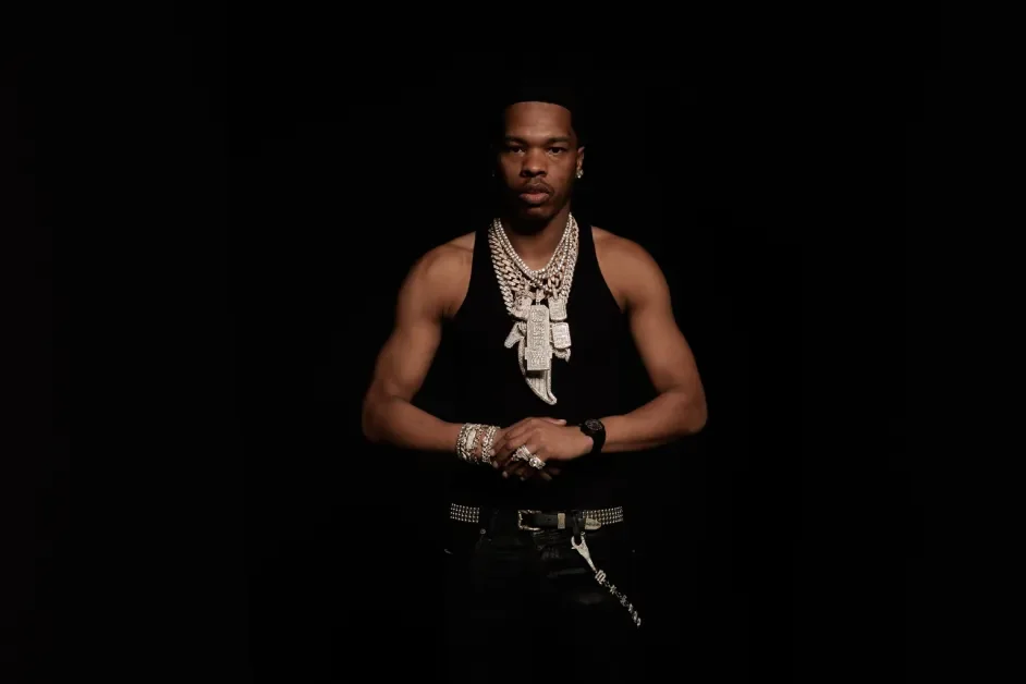 Young man wearing a black tank top and multiple silver jewelry, including necklaces, bracelets, rings, and a watch, standing against a black background.