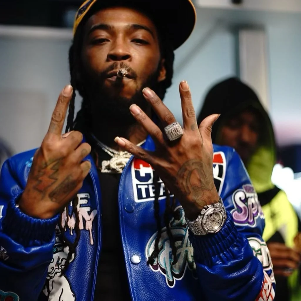 A man with tattoos on his hands and neck, wearing a blue racing jacket with various patches, a large diamond ring, a watch, and a black cap tilted sideways, smokes a cigarette, and makes a hand gesture with both hands.