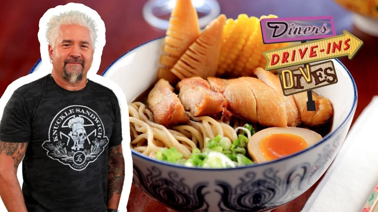 A man with gray hair and tattoos wearing a black T-shirt with white text and a skull logo stands next to a bowl of ramen with egg, pork, green onions, bamboo shoots, and corn, overlaid with colorful signs reading "Diners," "Drive-Ins," and "Dives."