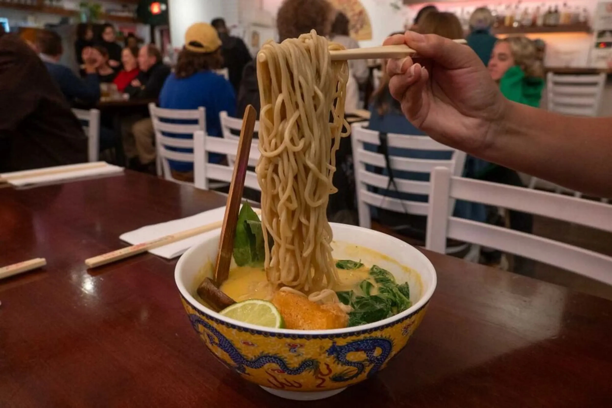 A person holding chopsticks lifting noodles from a bowl of ramen with vegetables and lime at a busy restaurant.