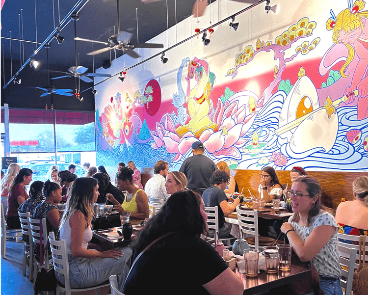 Busy restaurant with people dining at tables, large colorful mural of a dragon and mythological figures on the wall.