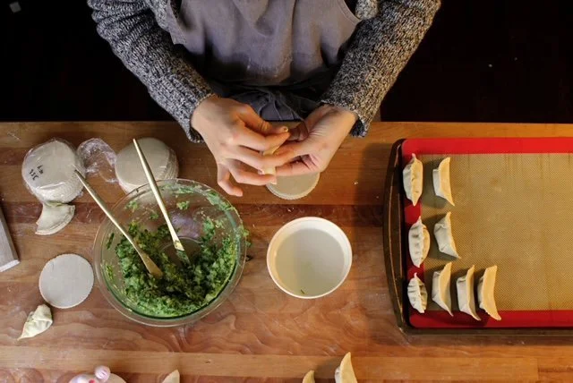 A person preparing dumplings with a tray of uncooked dumplings, a bowl of filling, and a bowl of water on a wooden table.
