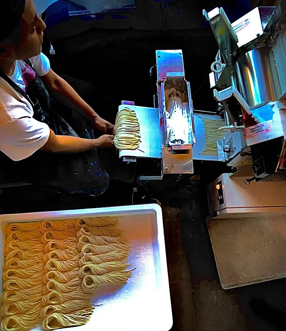 Person operating a pasta machine, making fresh noodles with rolled dough, with neatly arranged curls on a tray in the foreground.